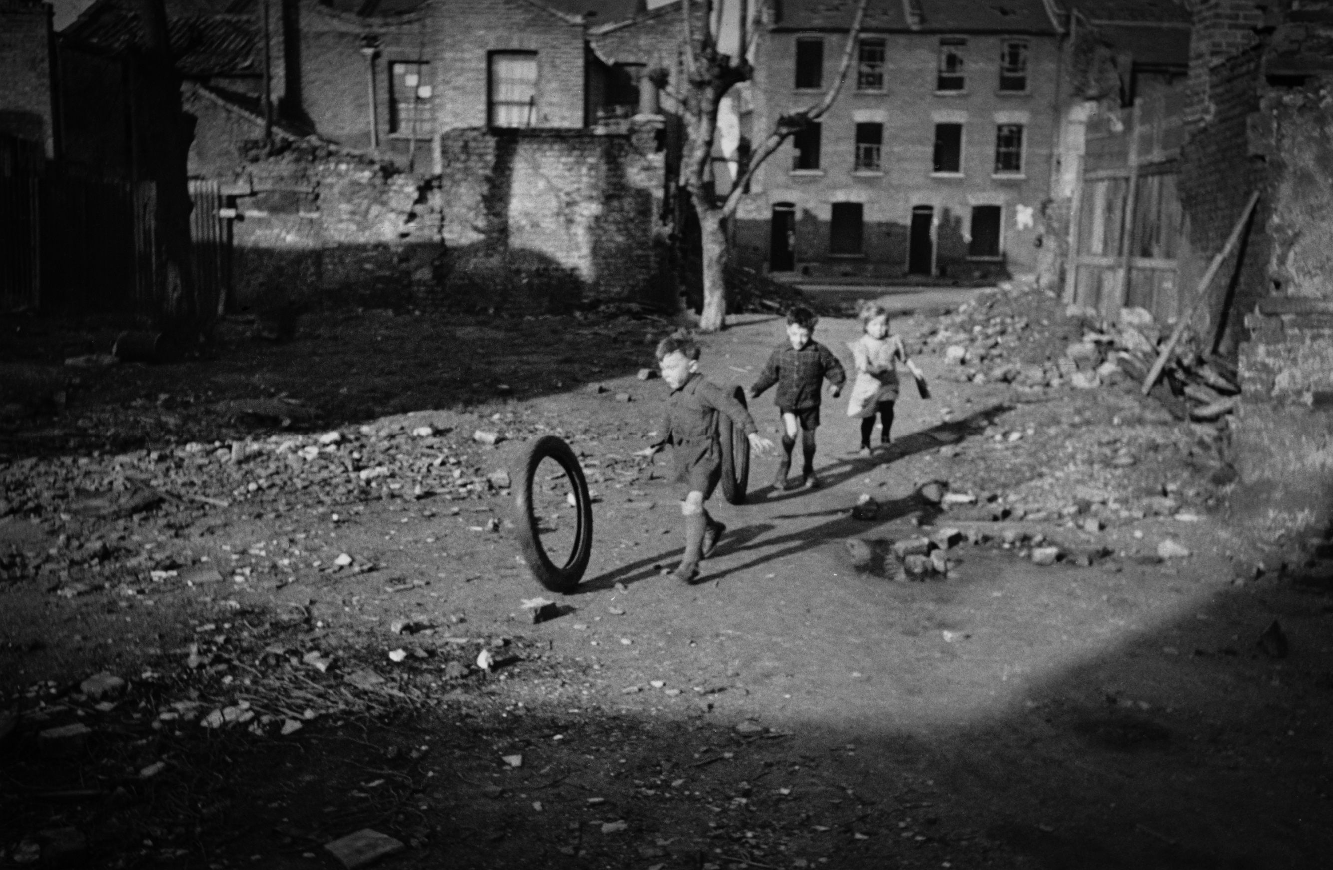 Children in an urban setting play with a tyre in a rubble-strewn street surrounded by old brick buildings.
