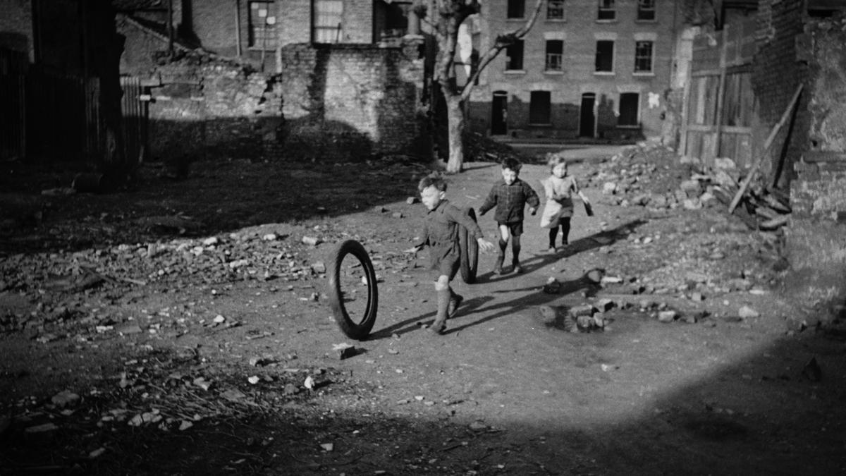 Children in an urban setting play with a tyre in a rubble-strewn street surrounded by old brick buildings.