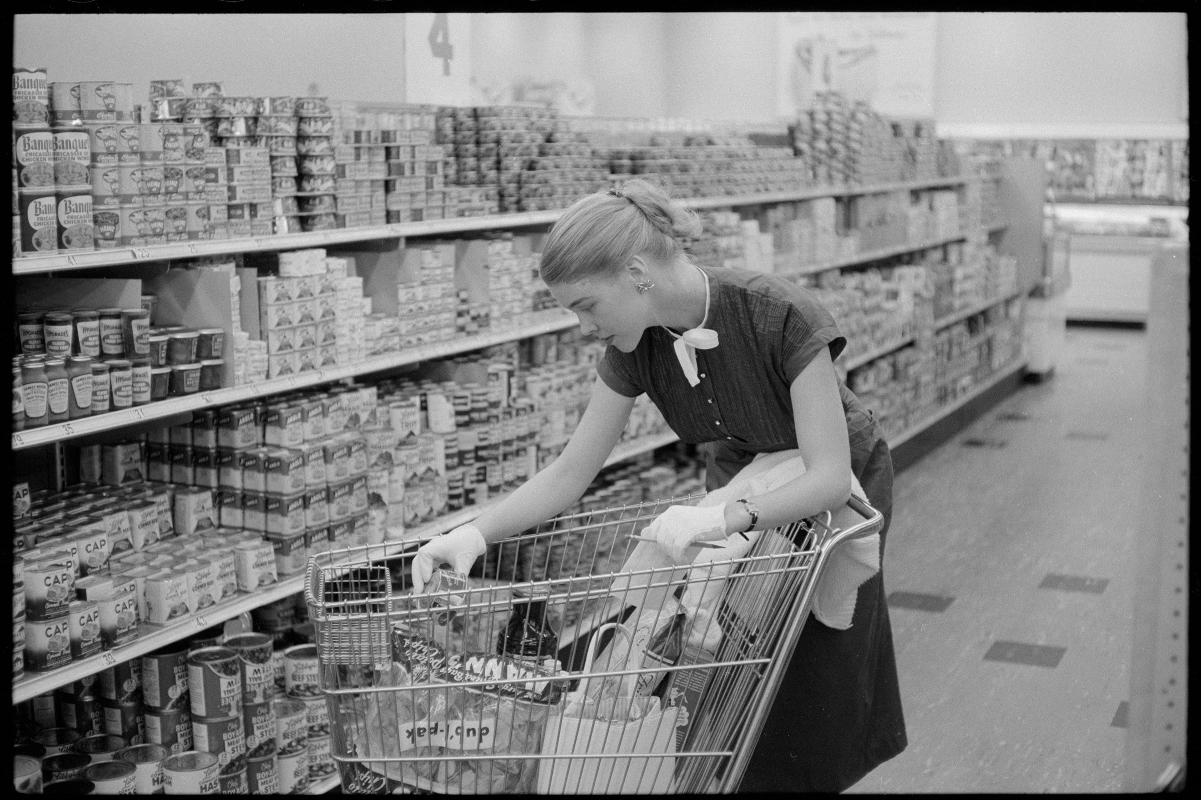 Black and white photo of a woman shopping in a supermarket aisle, placing items in a trolley filled with groceries.