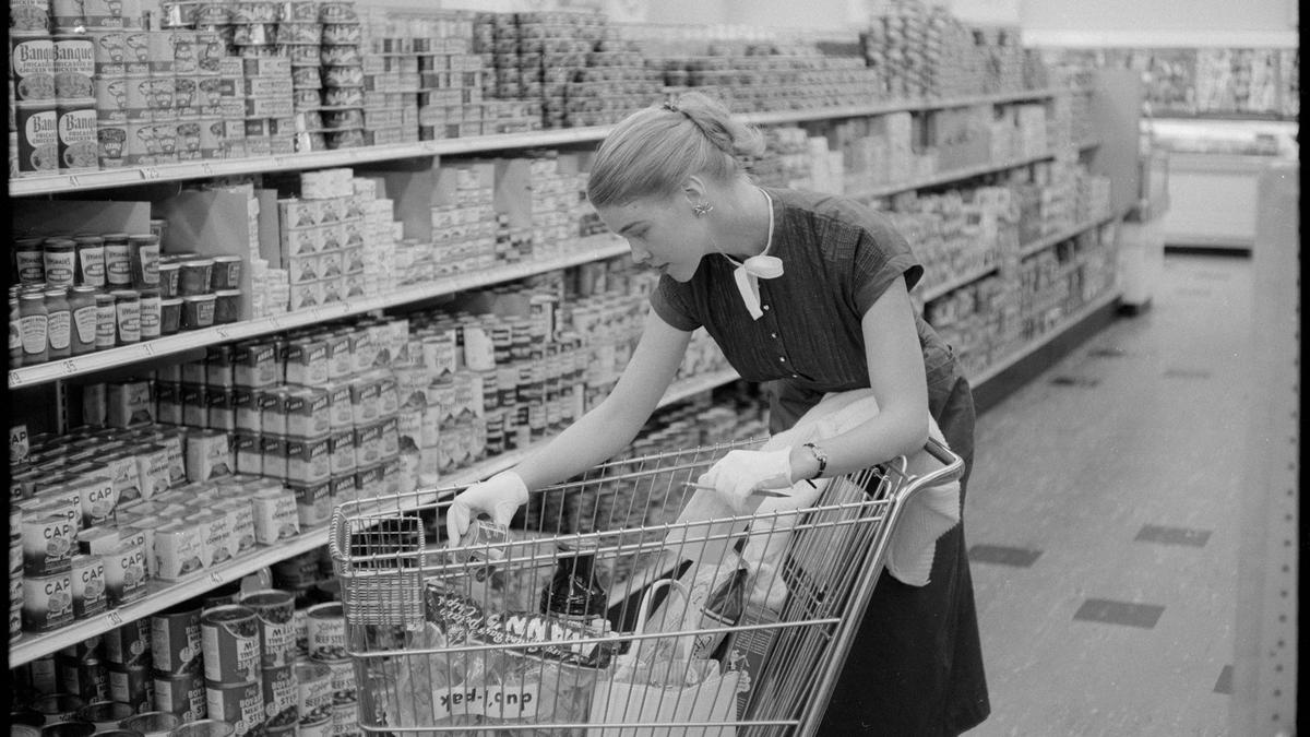 Black and white photo of a woman shopping in a supermarket aisle, placing items in a trolley filled with groceries.