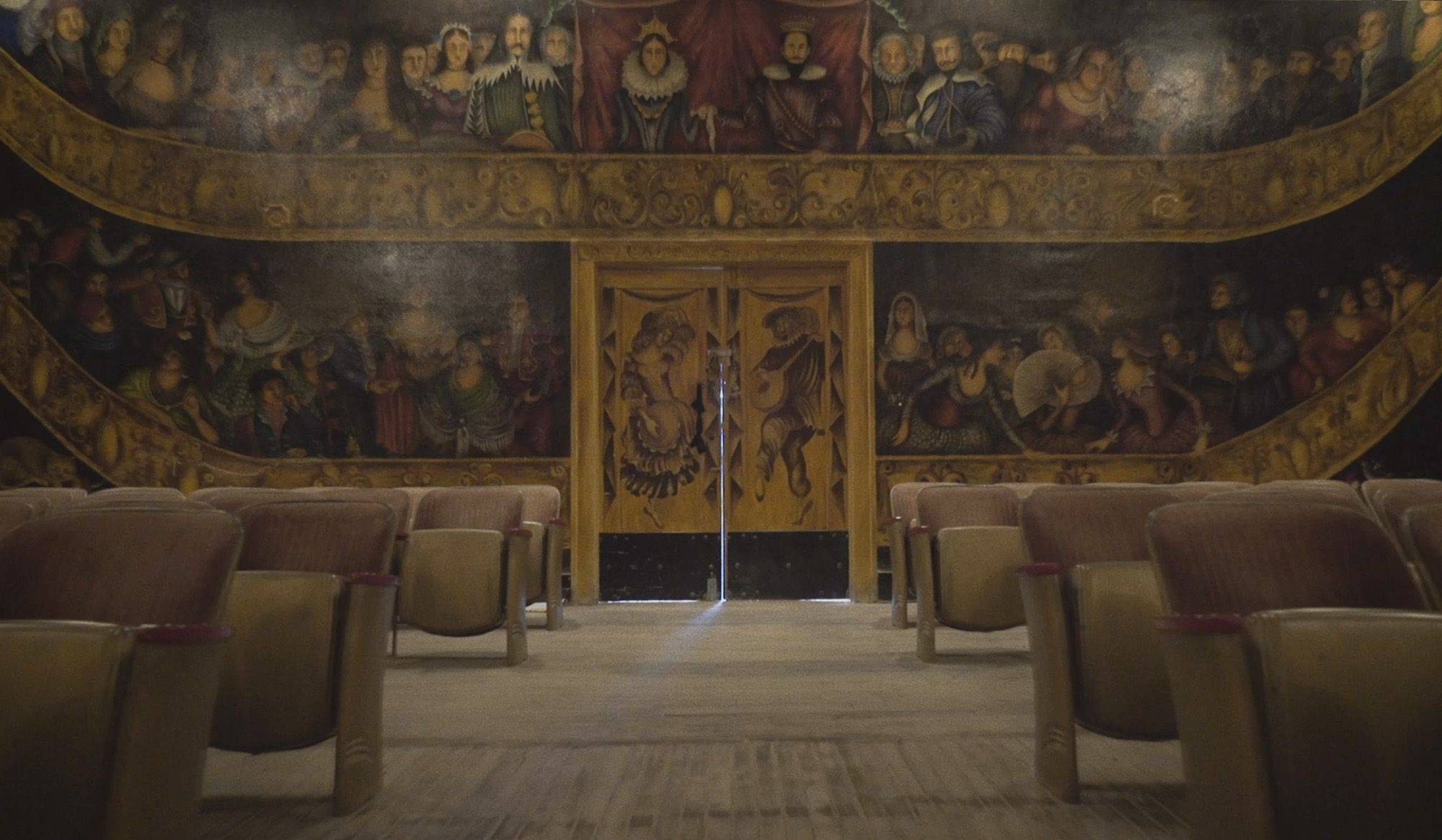 An old theatre with rows of empty seats. The walls are painted with a detailed mural of people in period clothing.