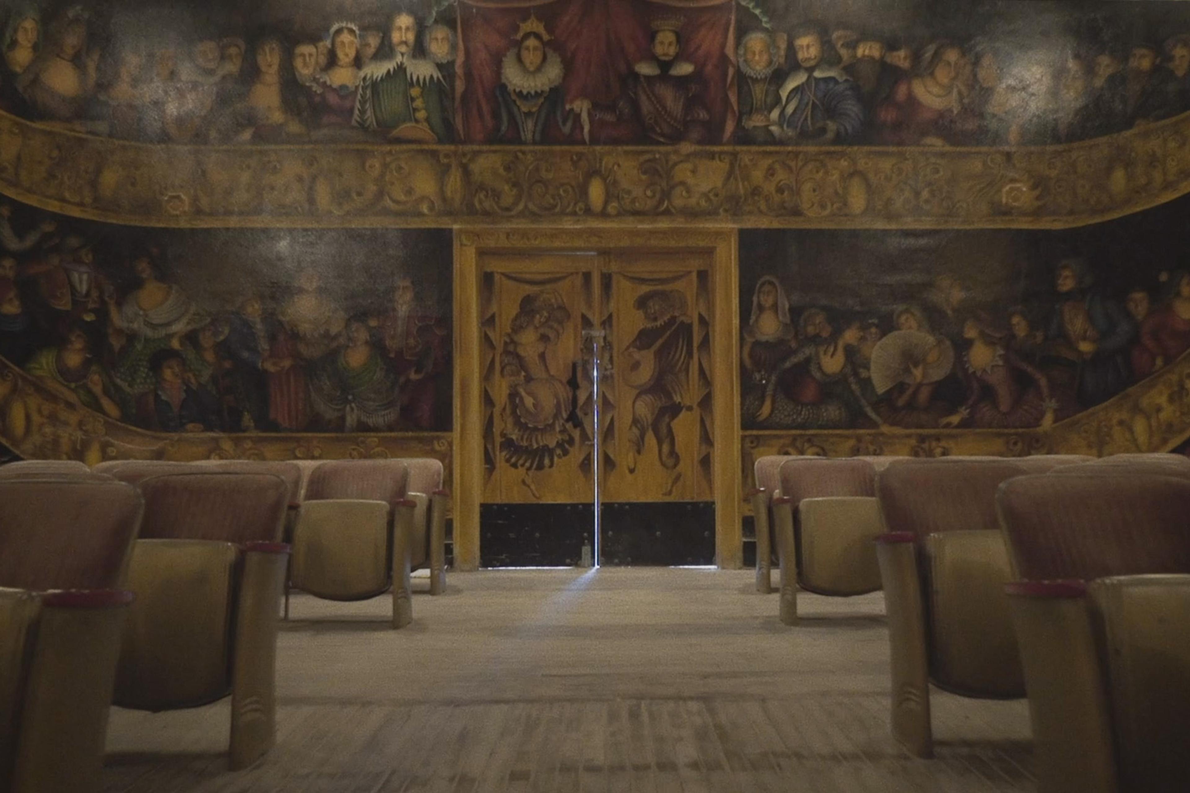 An old theatre with rows of empty seats. The walls are painted with a detailed mural of people in period clothing.