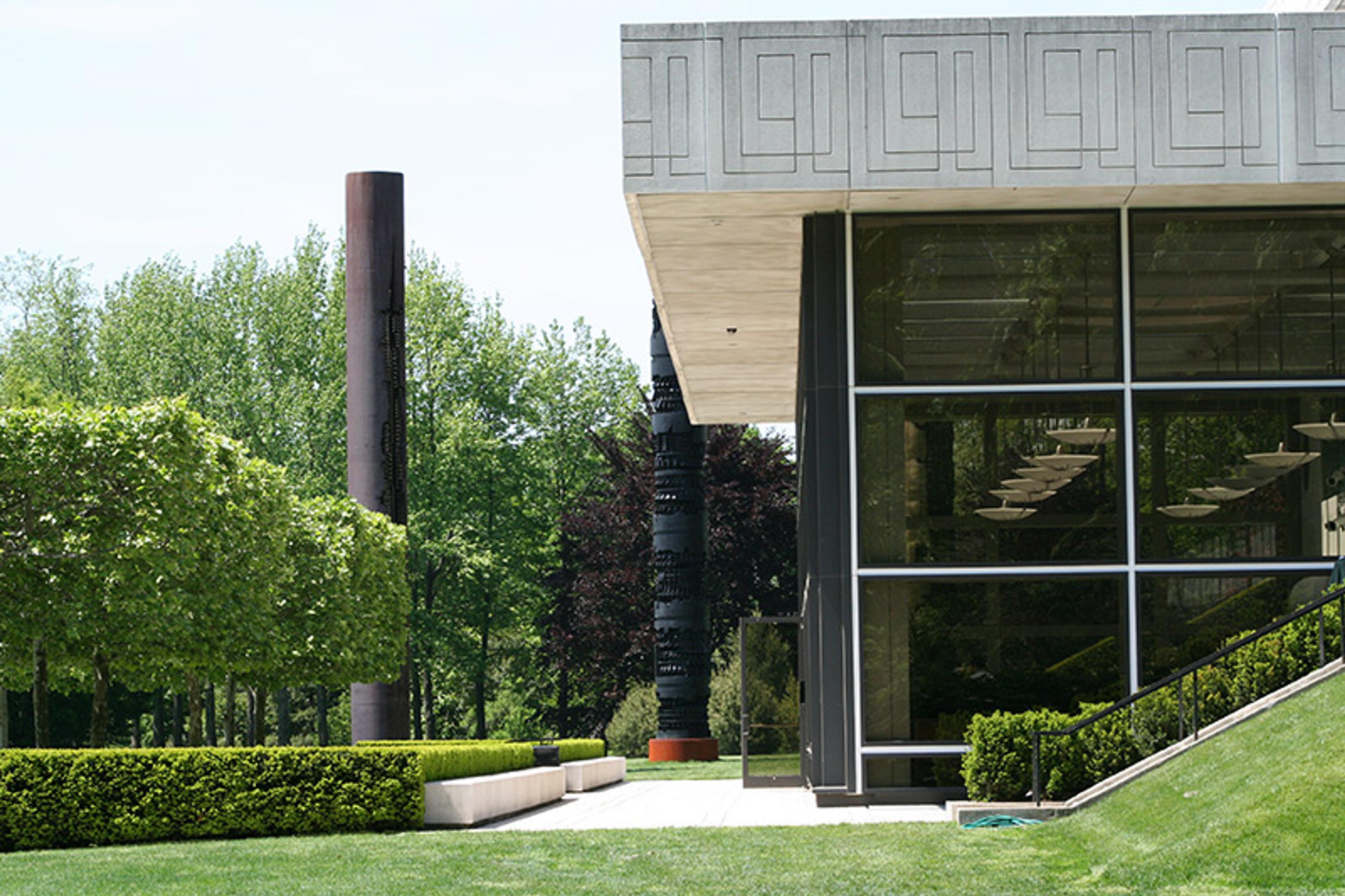 A modern building with large windows, surrounded by green trees and bushes, and featuring tall cylindrical sculptures.