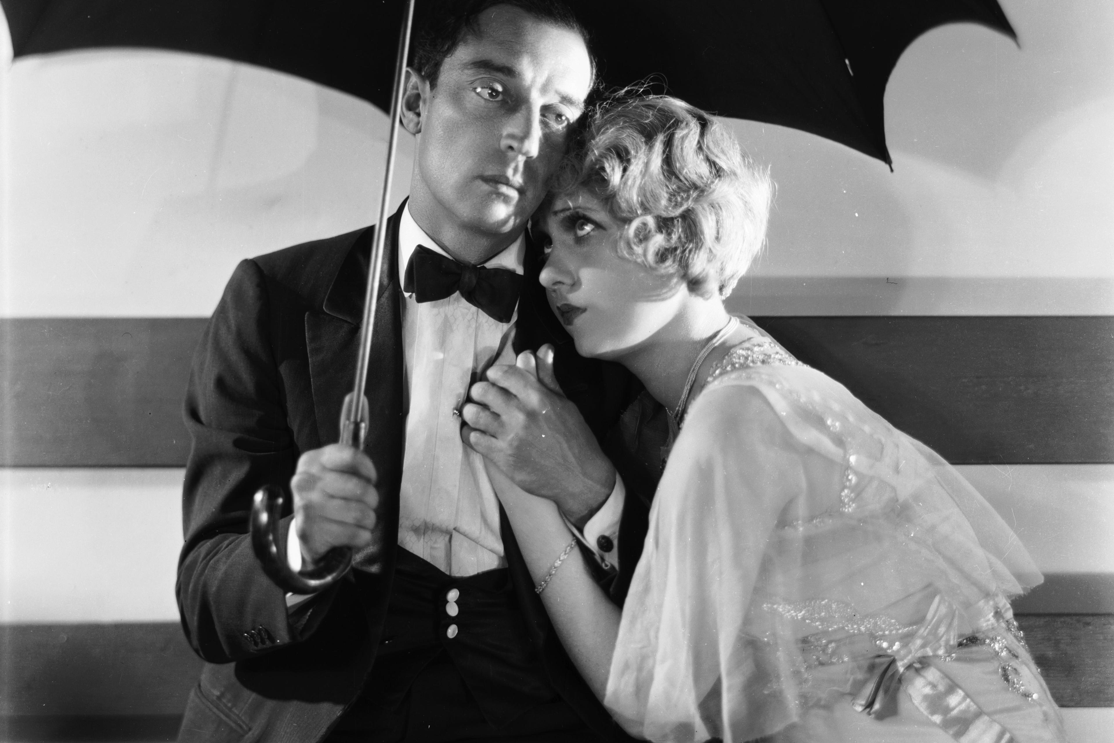 Black and white photo of a man and woman sitting under an umbrella; the woman leans on the man’s shoulder.