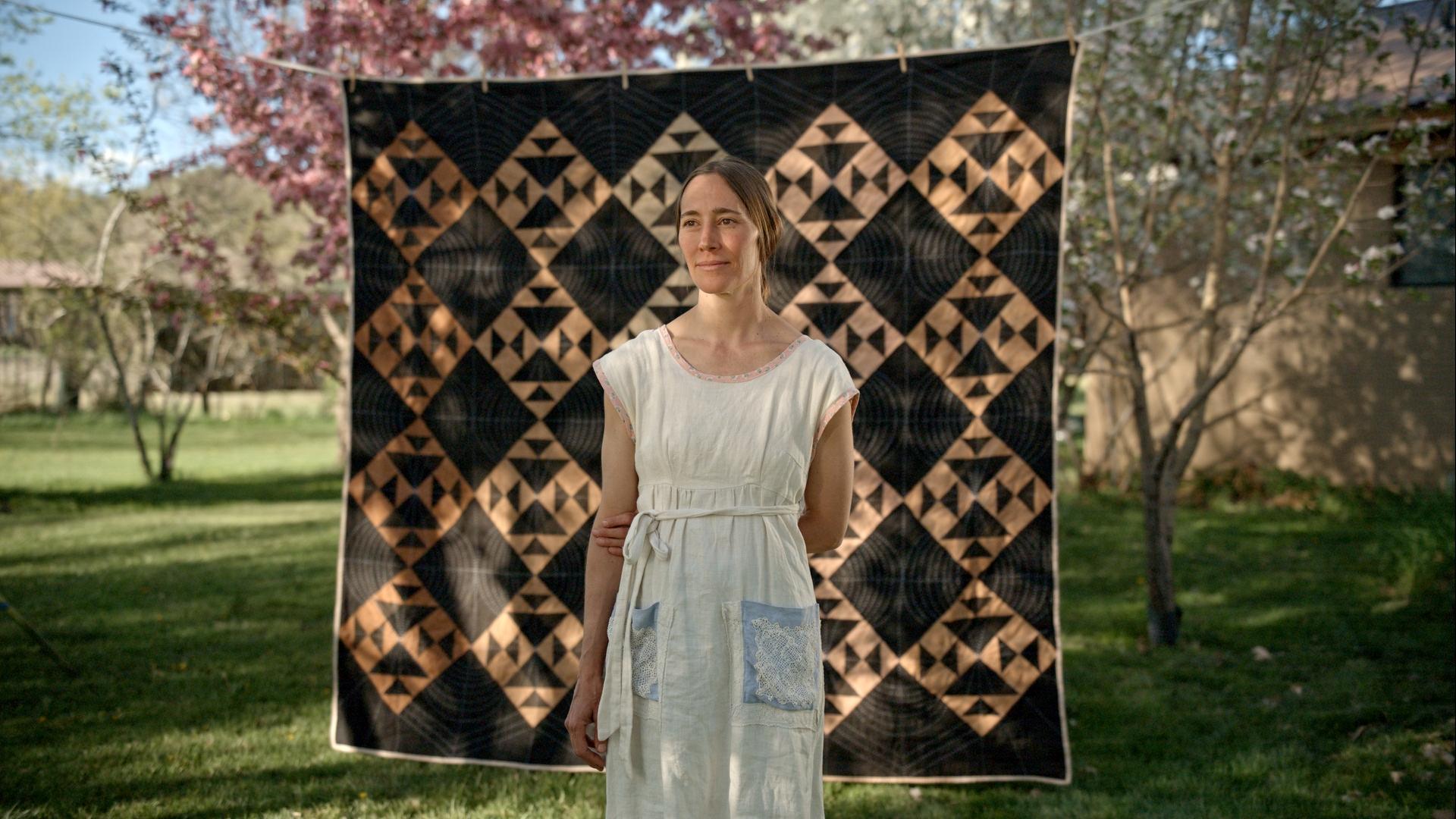 Woman in a white dress standing in front of a geometric quilt in a garden setting with trees in the background.