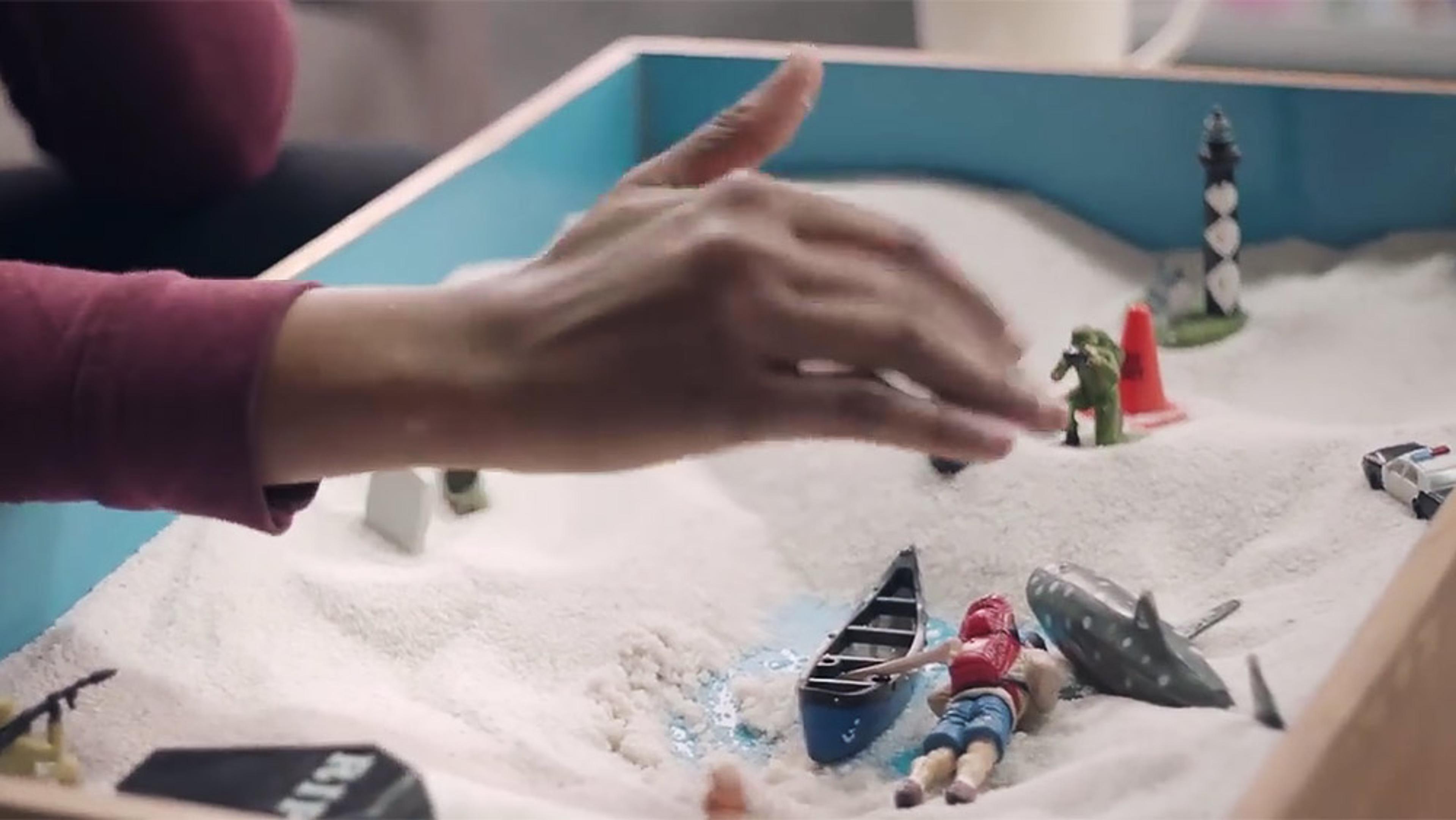 A hand arranging miniature figures in a sandbox. The scene includes a canoe, a person, a shark, and a lighthouse.