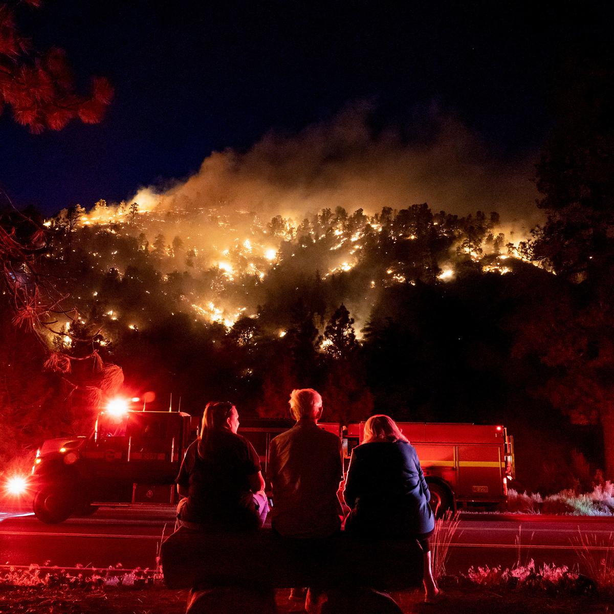 Three people sit on a bench facing a forest fire on a hill at night, with a fire truck nearby. The flames illuminate the trees, creating a dramatic and intense scene.