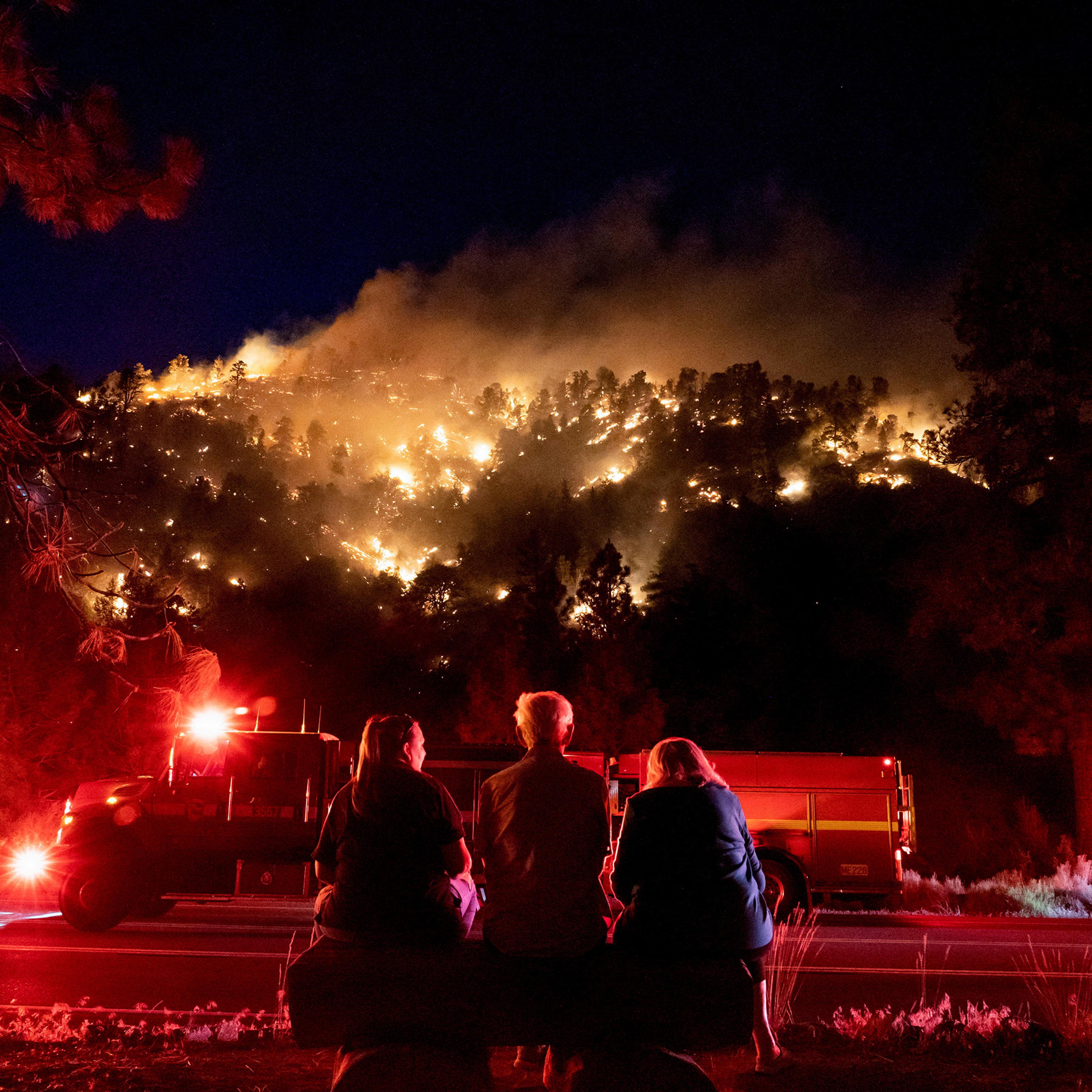 Three people sit on a bench facing a forest fire on a hill at night, with a fire truck parked nearby. The flames illuminate the trees, creating a dramatic and intense scene.