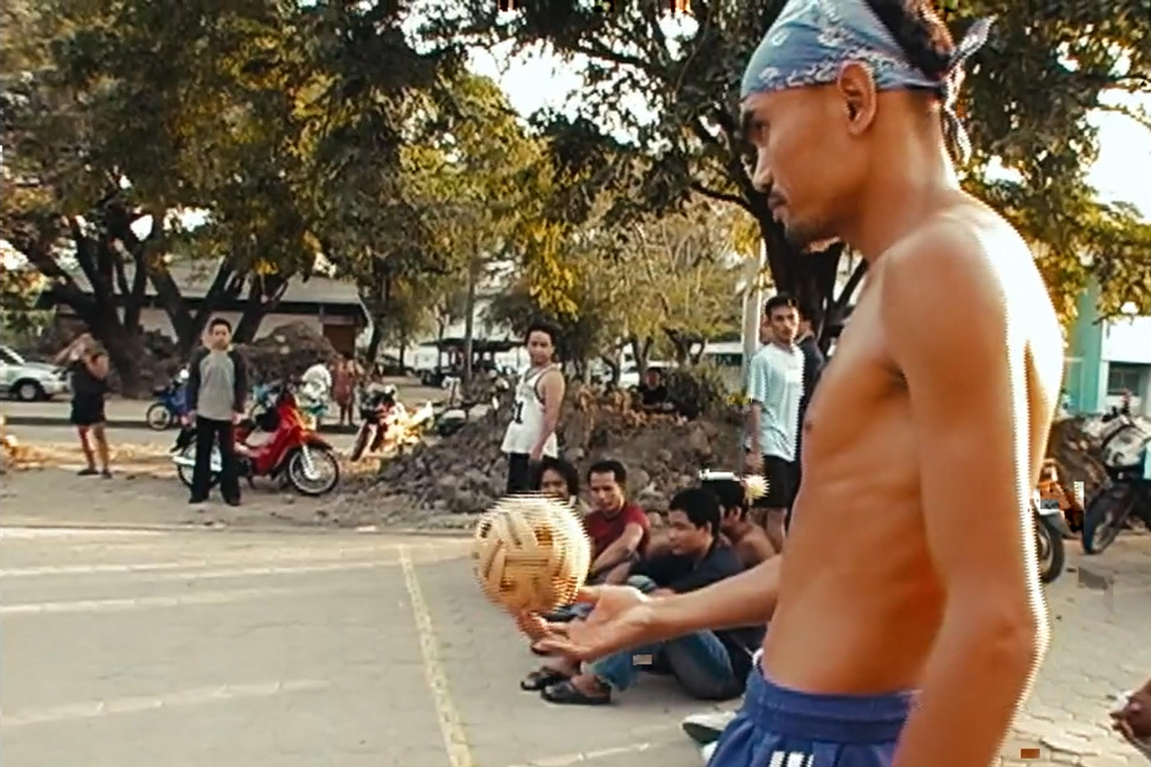 Photo of a shirtless man balancing a woven ball on his hand, with onlookers seated on a street lined with trees.