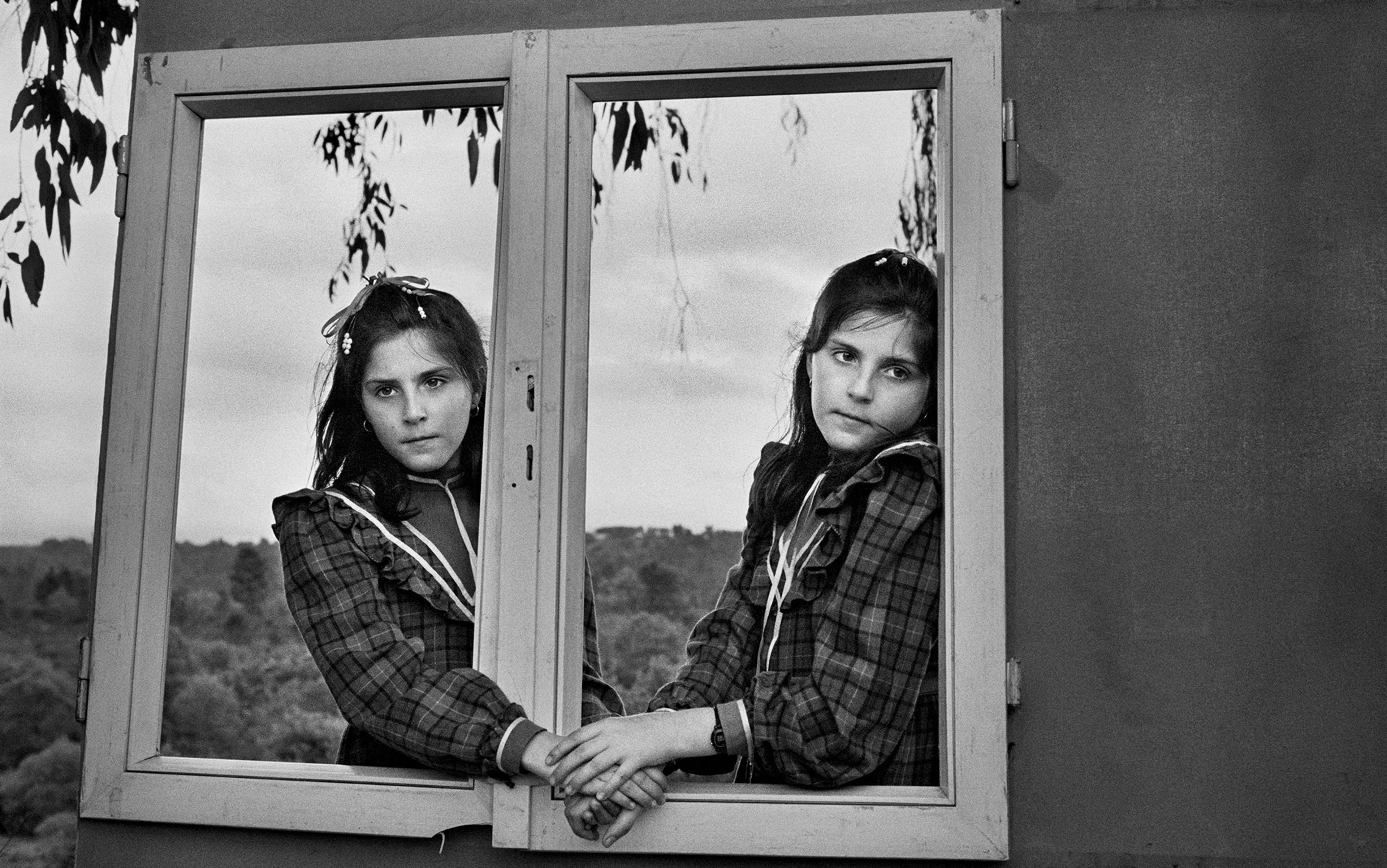 Two dark haired twin girls in tartan dresses look through an unglazed window frame