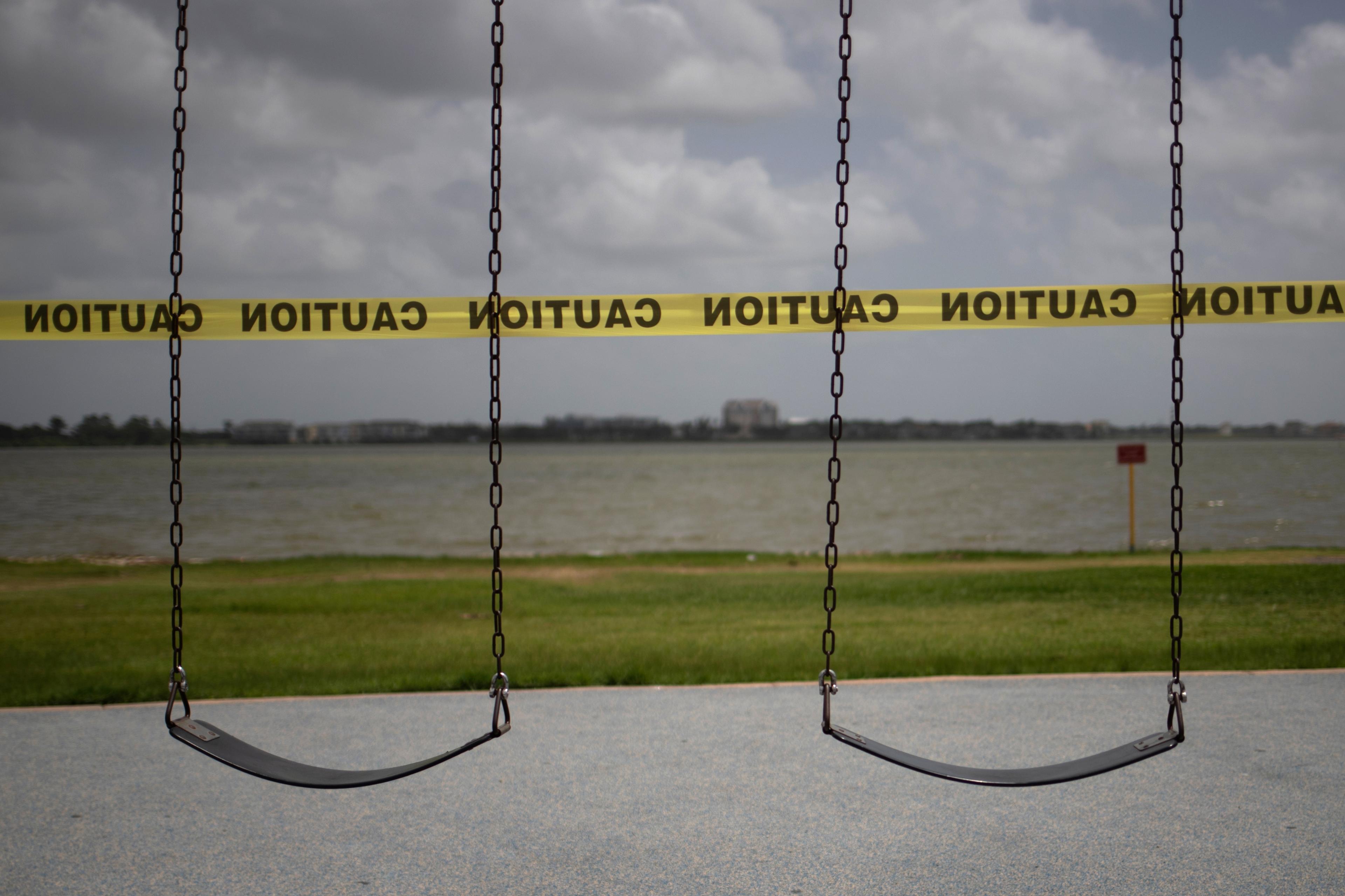 Photo of two empty swings by a lake, cordoned off with yellow caution tape against a cloudy sky.