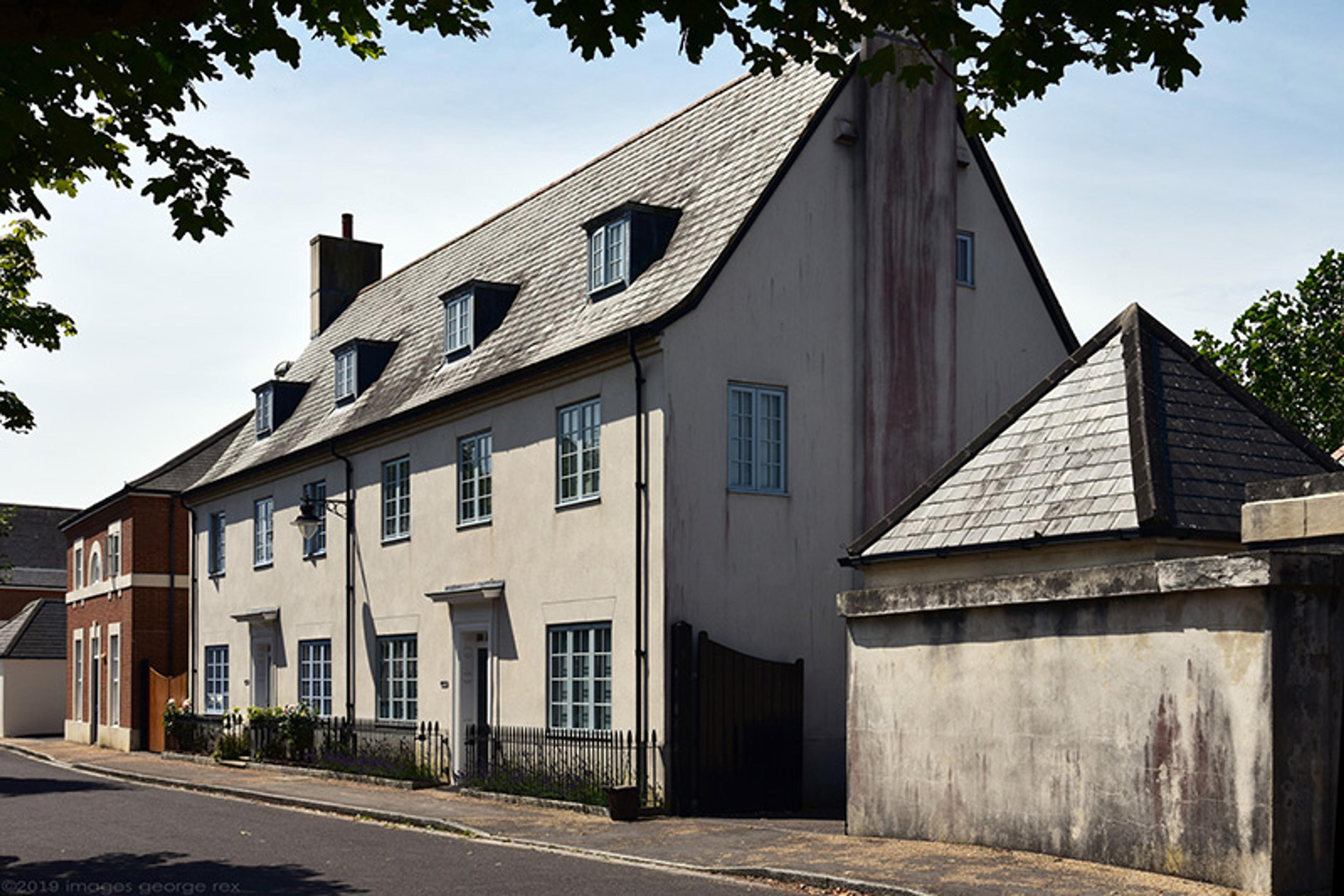 A white three-storey house with a grey roof on a quiet street, surrounded by trees and neighbouring buildings.