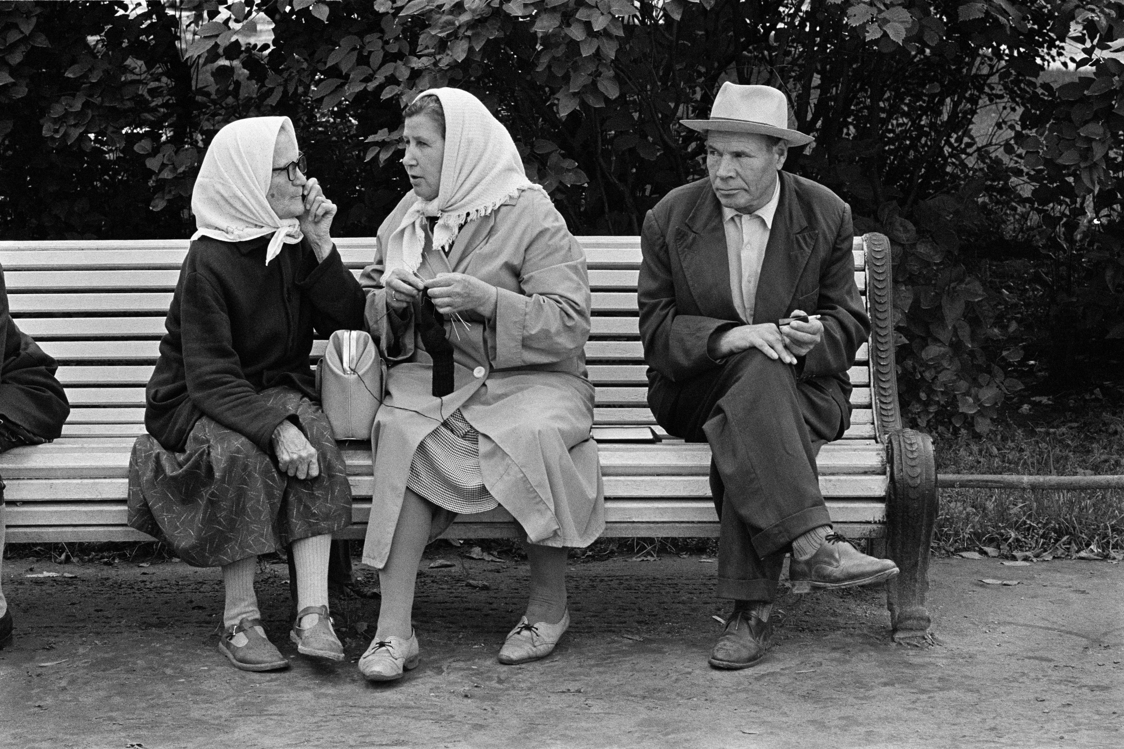 Black and white photo of three elderly people on a bench; two women in headscarves and a man in a hat; they appear engaged.
