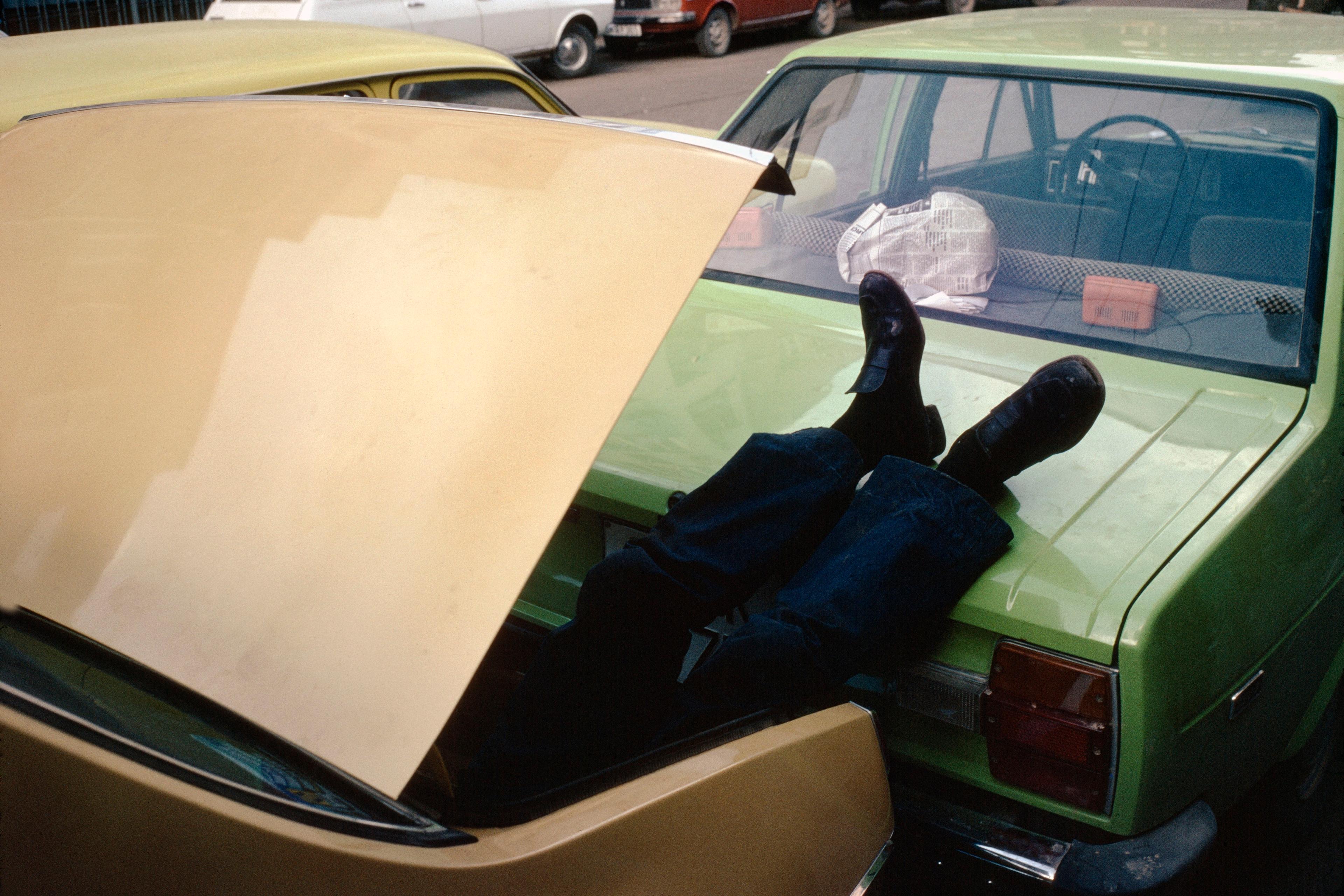 Photo of a person lying in a car boot with legs sticking out, parked cars in background.