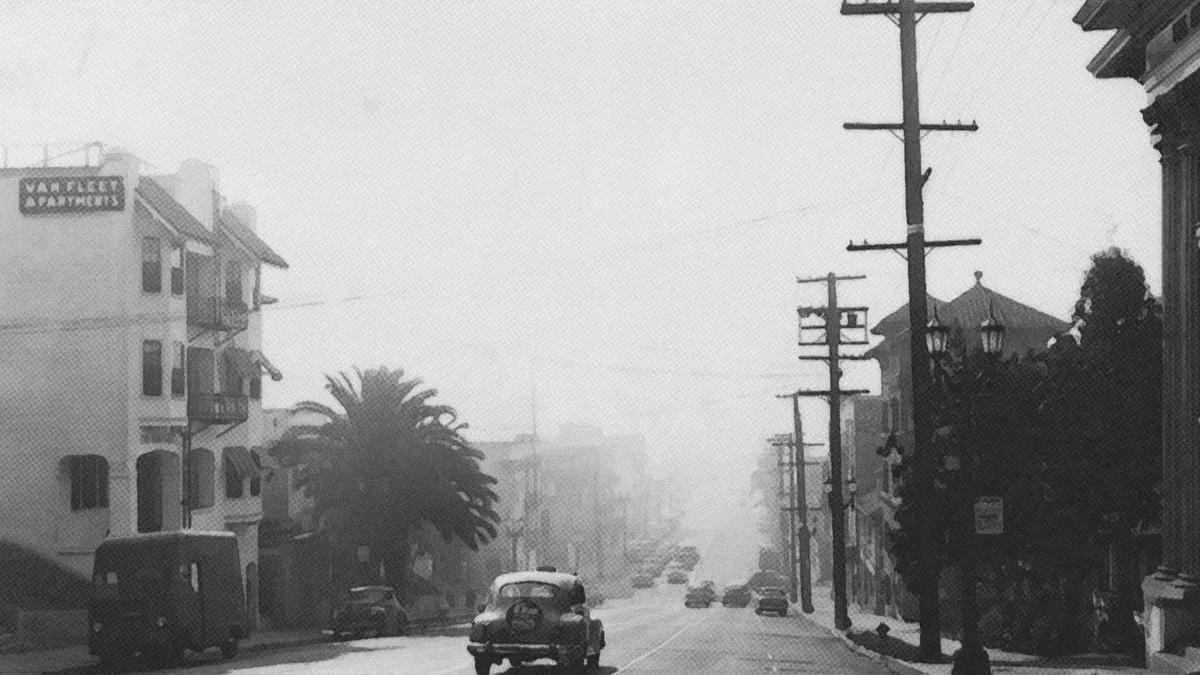 Black and white photo of a vintage car on a foggy city street lined with buildings and palm trees.