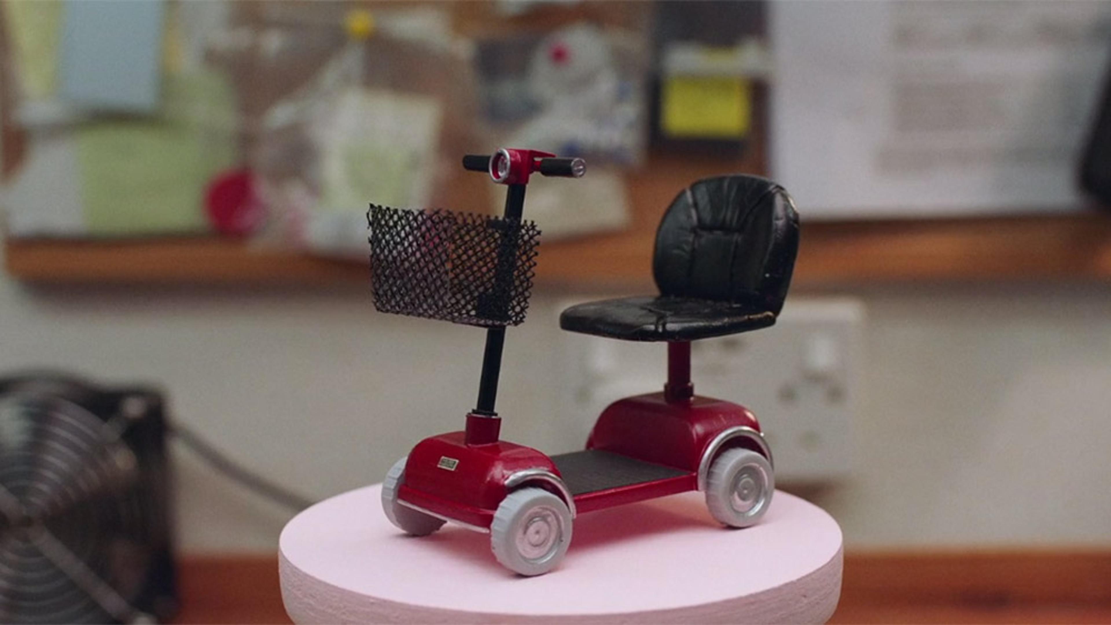A small red mobility scooter model with a black seat and basket on display, with a blurred bulletin board in the background.