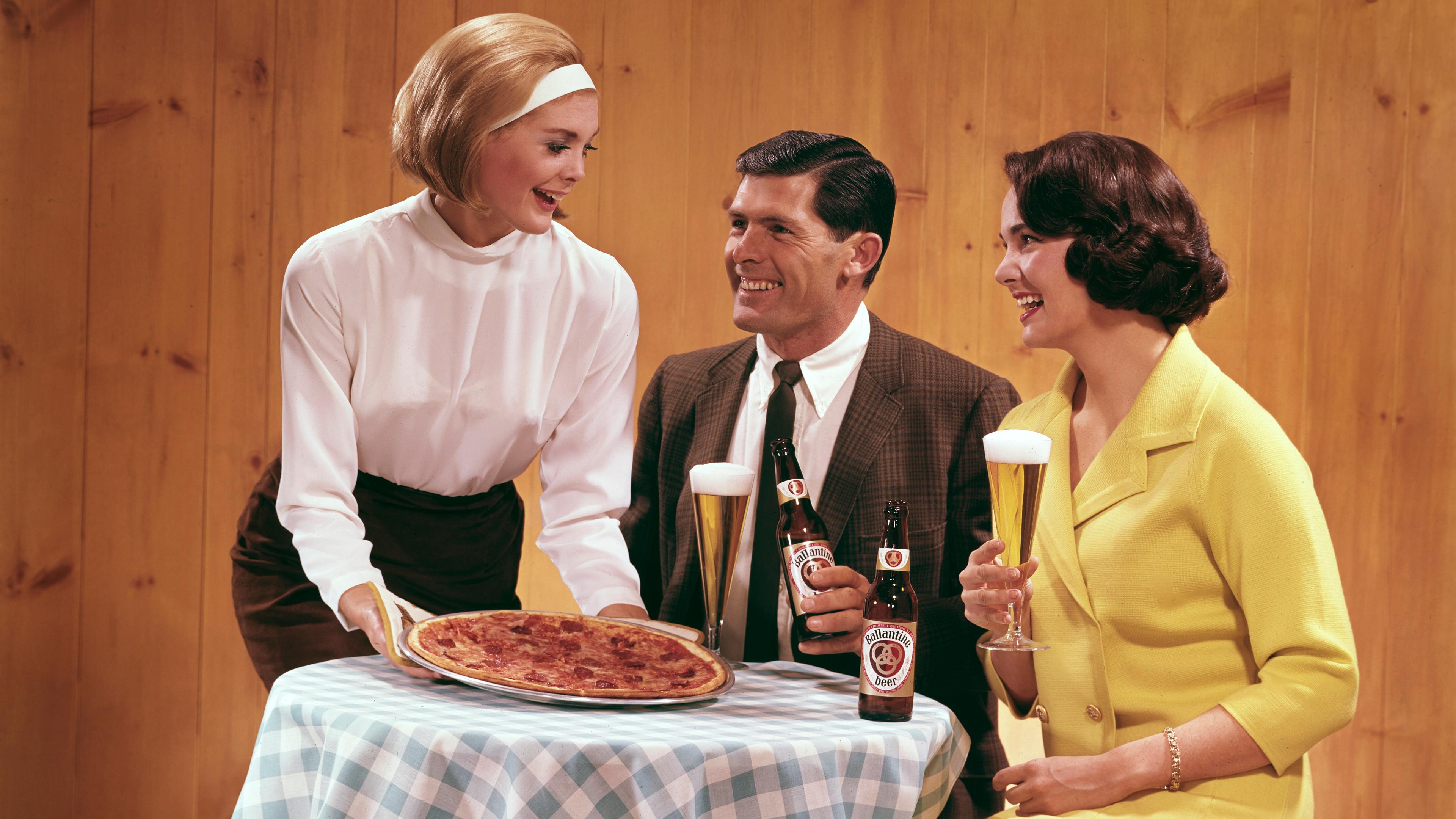 Retro photo of a waitress serving pizza to a smiling couple seated at a table with beer and glasses.