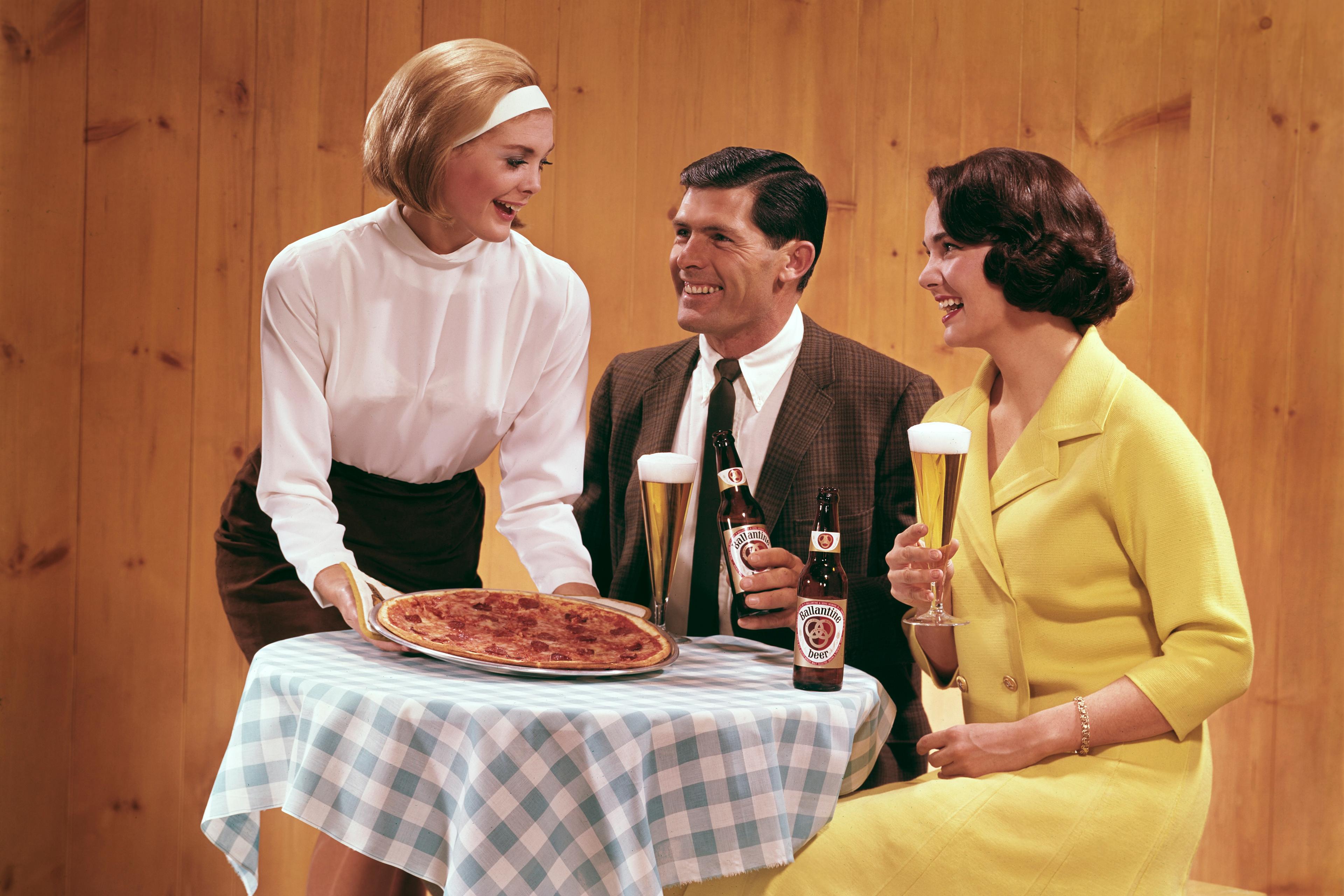 Retro photo of a waitress serving pizza to a smiling couple seated at a table with beer and glasses.