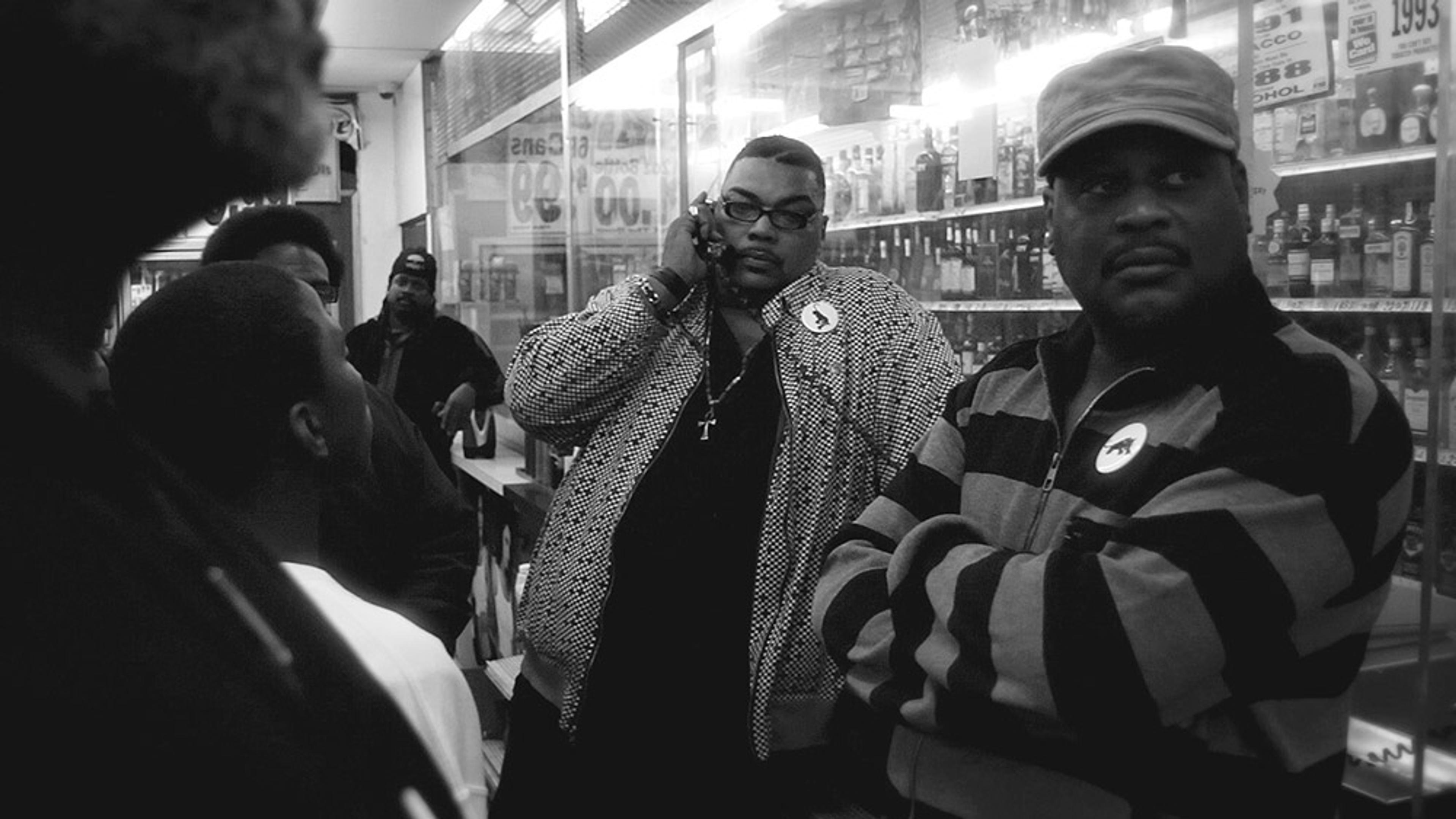 Black and white photo of a group of men in a liquor store, one man is talking on a mobile phone with arms crossed.