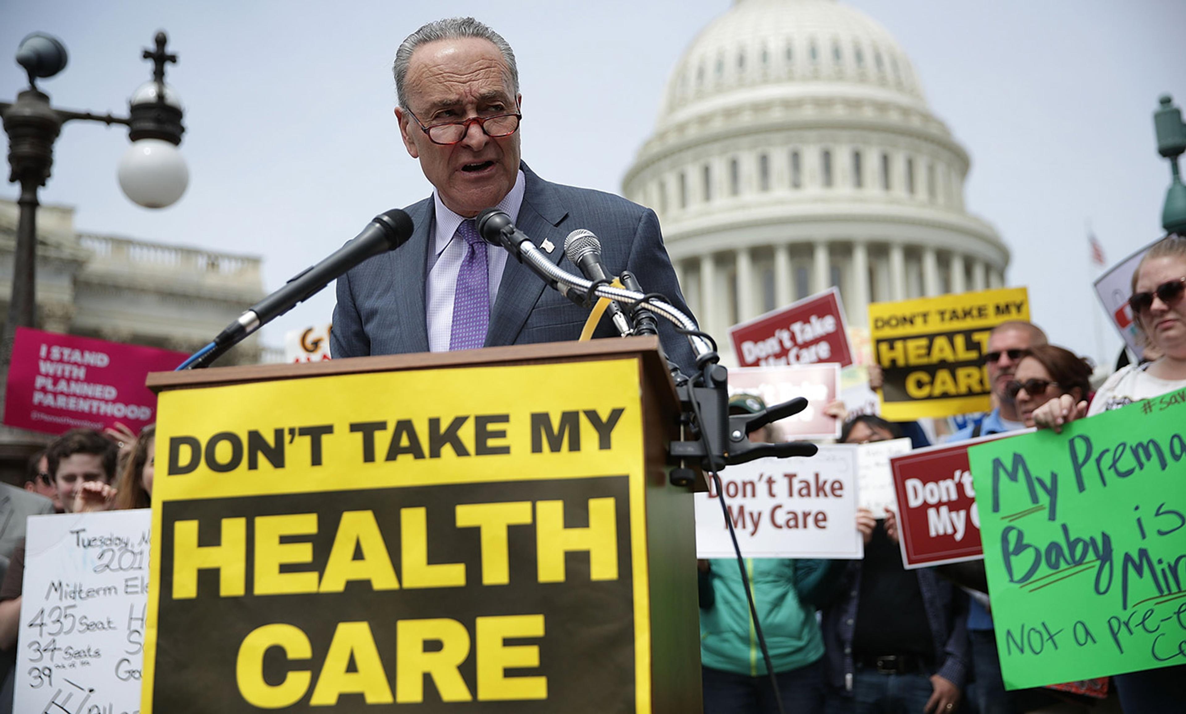 A man in a suit and tie at a podium in front of the US Capitol with a ‘Don’t Take My Health Care’ sign, surrounded by protesters holding similar signs.