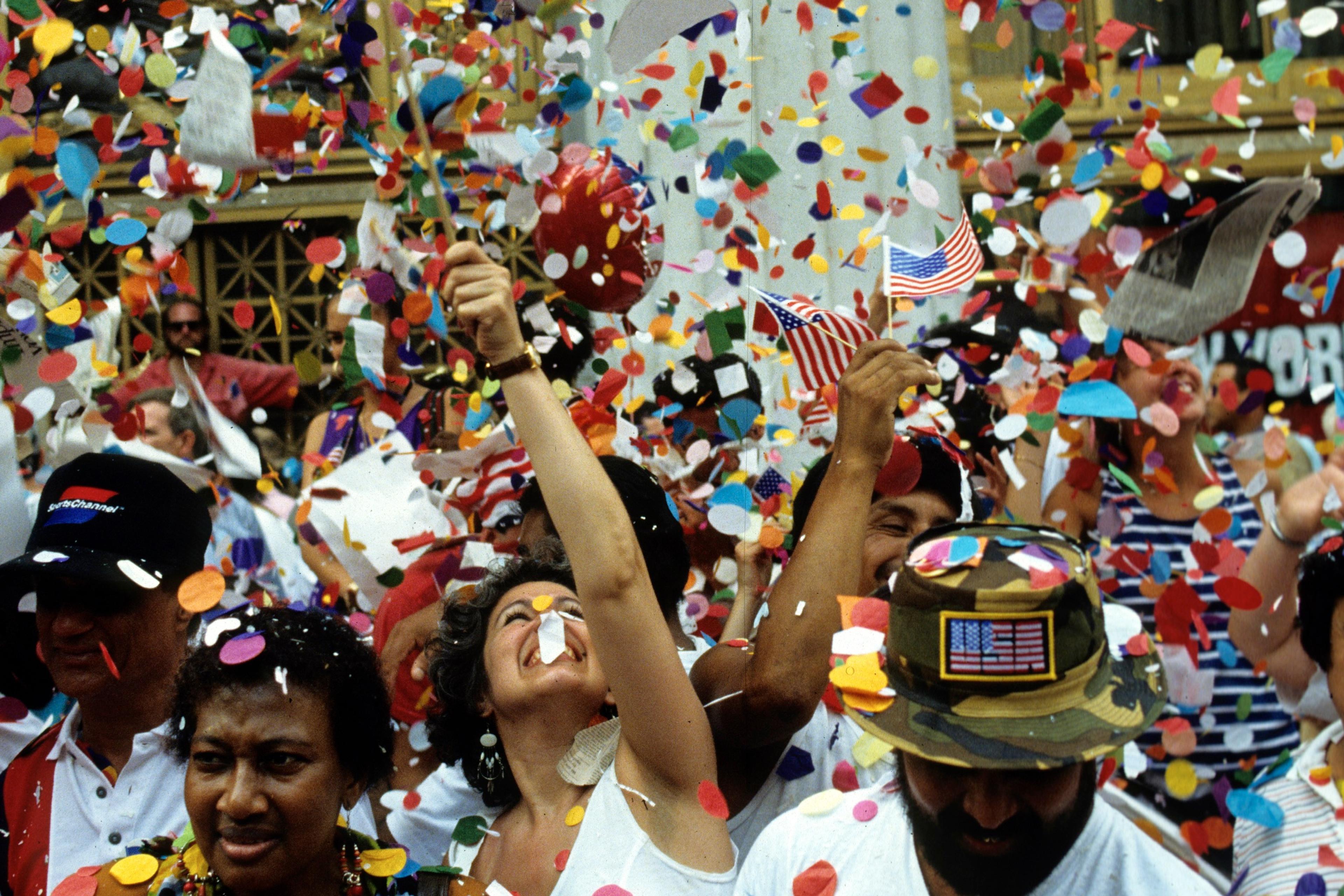 Photo of a vibrant street parade with people celebrating amid swirling confetti and waving American flags.