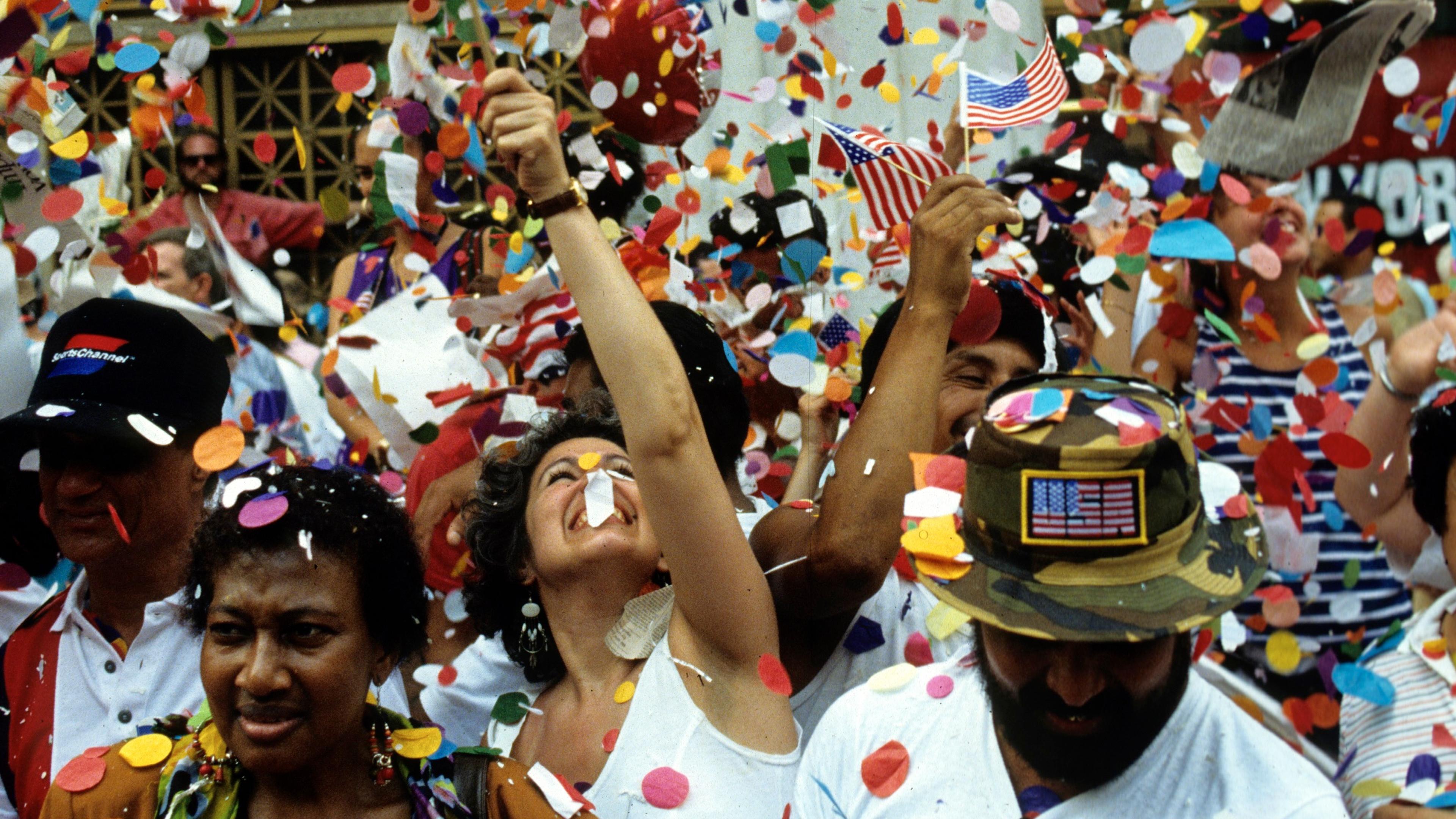 Photo of a vibrant street parade with people celebrating amid swirling confetti and waving American flags.