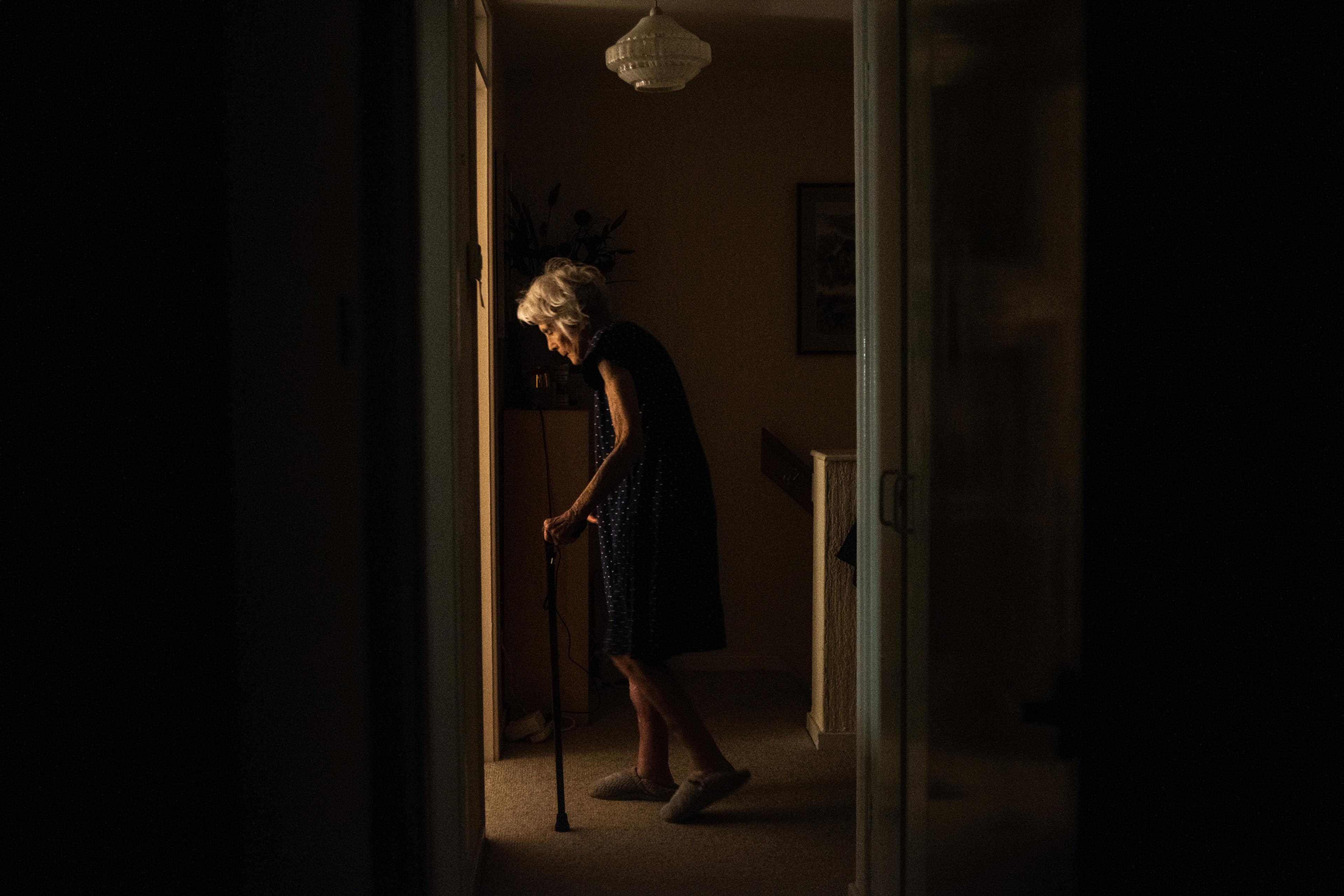 An elderly person walking with a cane in a dimly lit hallway, light from a room accentuates their silhouette.