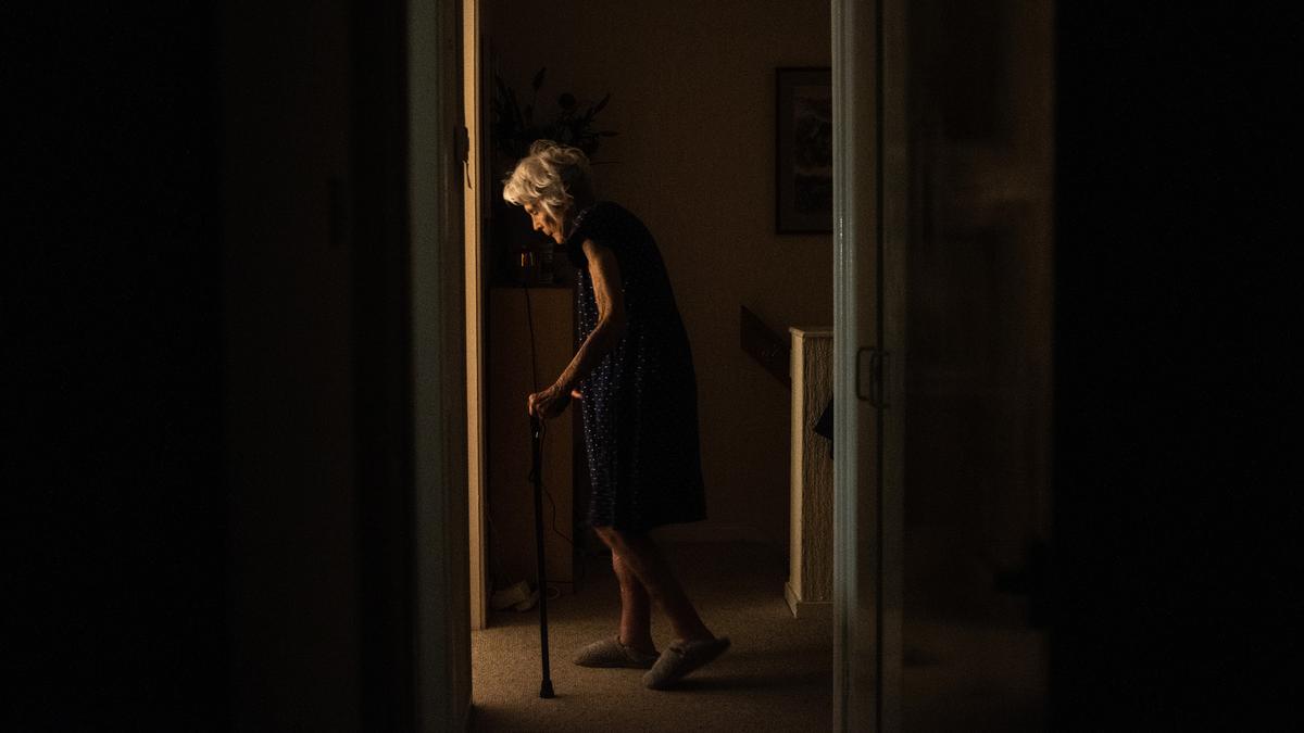 An elderly person walking with a cane in a dimly lit hallway, light from a room accentuates their silhouette.