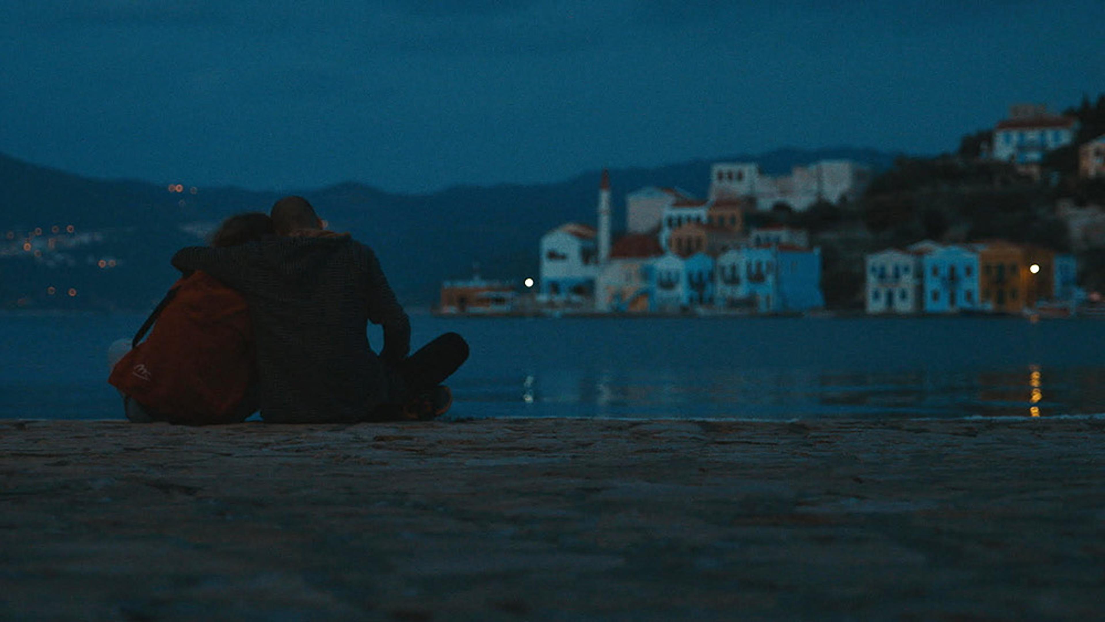 Two people embracing by the water at dusk with their backs to the camera, colourful buildings and mountains across the bay.