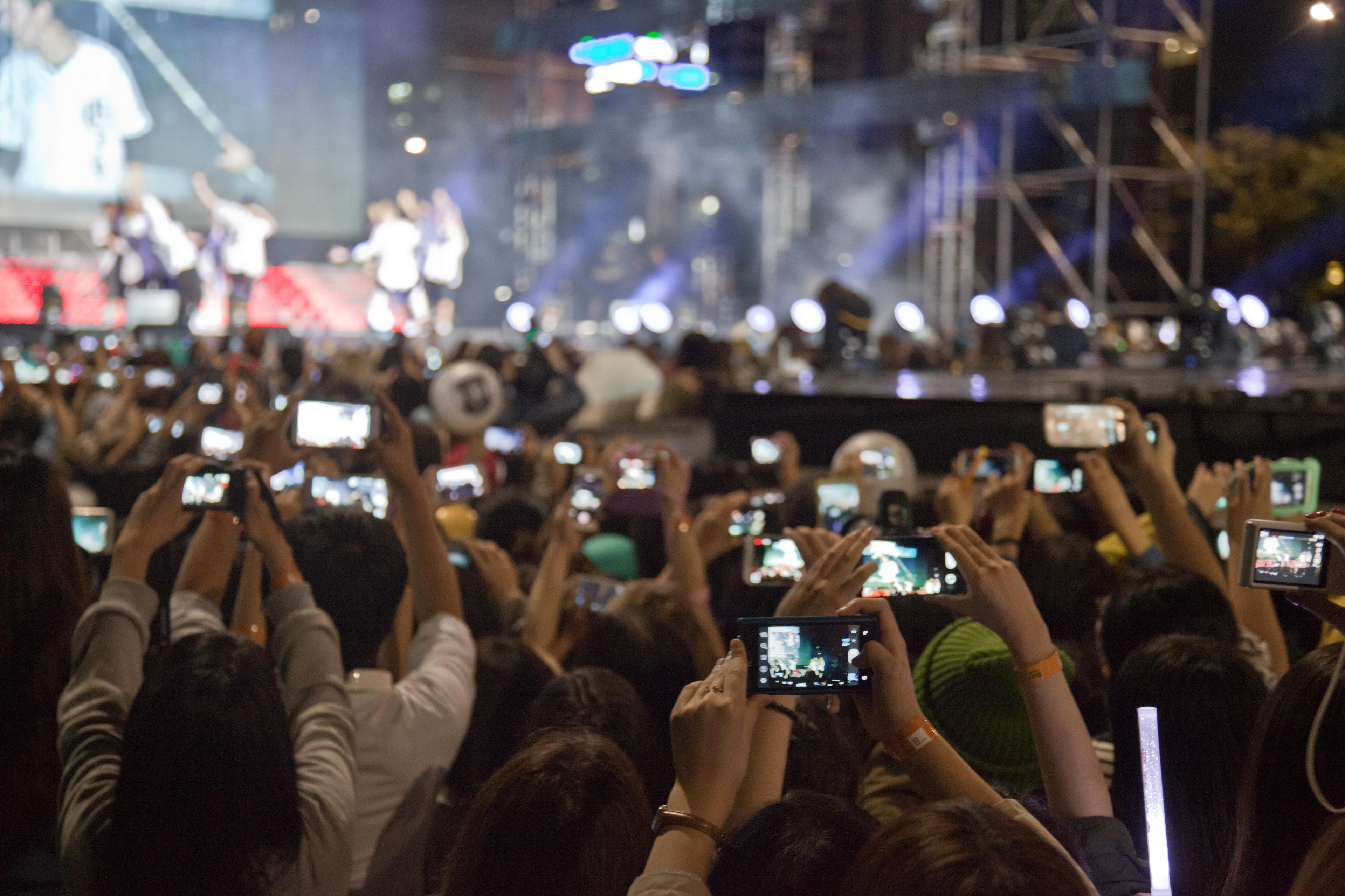 Photo of a concert audience holding up phones to record a performance on stage with bright lights in the background.