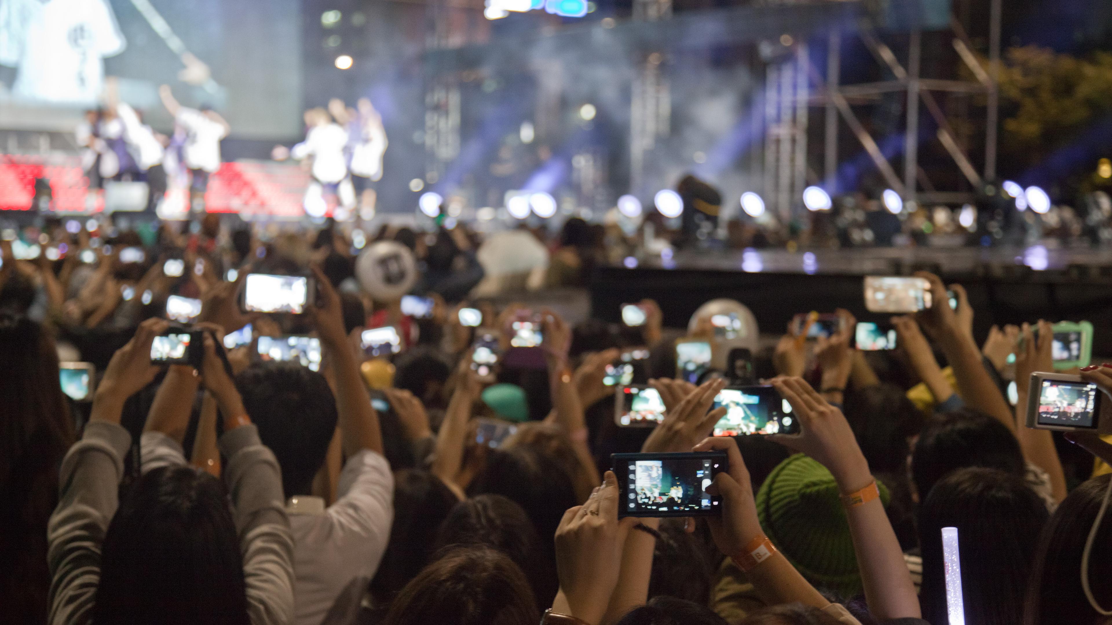 Photo of a concert audience holding up phones to record a performance on stage with bright lights in the background.