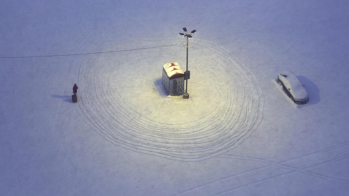 Aerial photo of a snow-covered bus stop with lights, car covered in snow and person walking with luggage, tracks in snow visible.