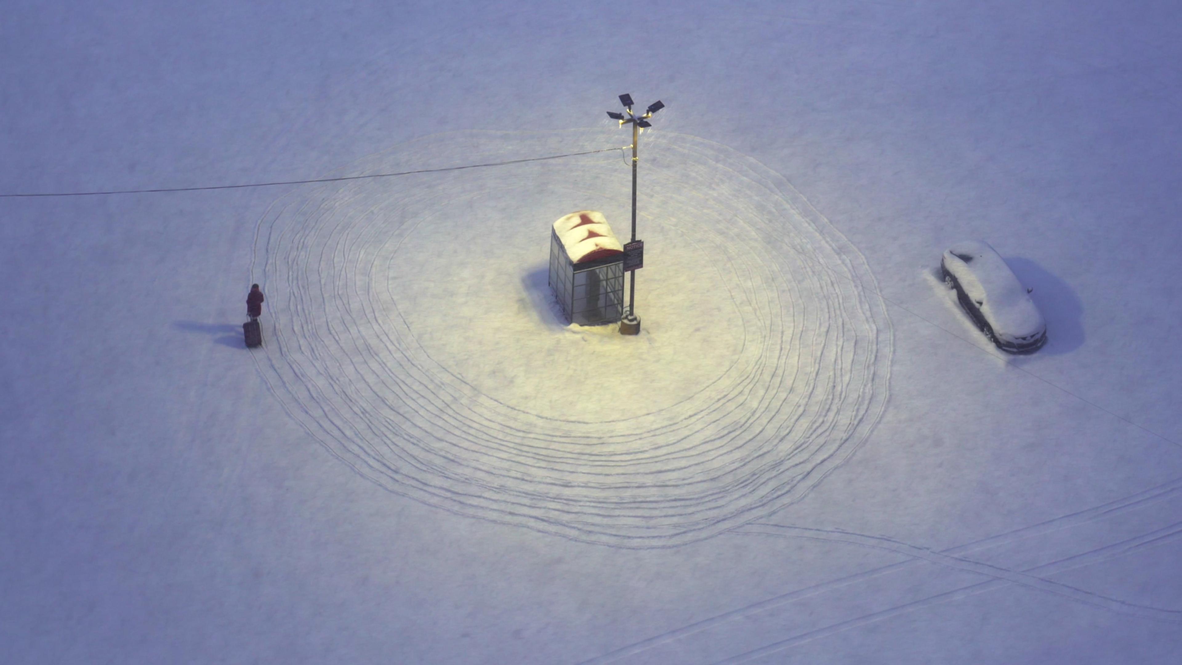 Aerial photo of a snow-covered bus stop with lights, car covered in snow and person walking with luggage, tracks in snow visible.