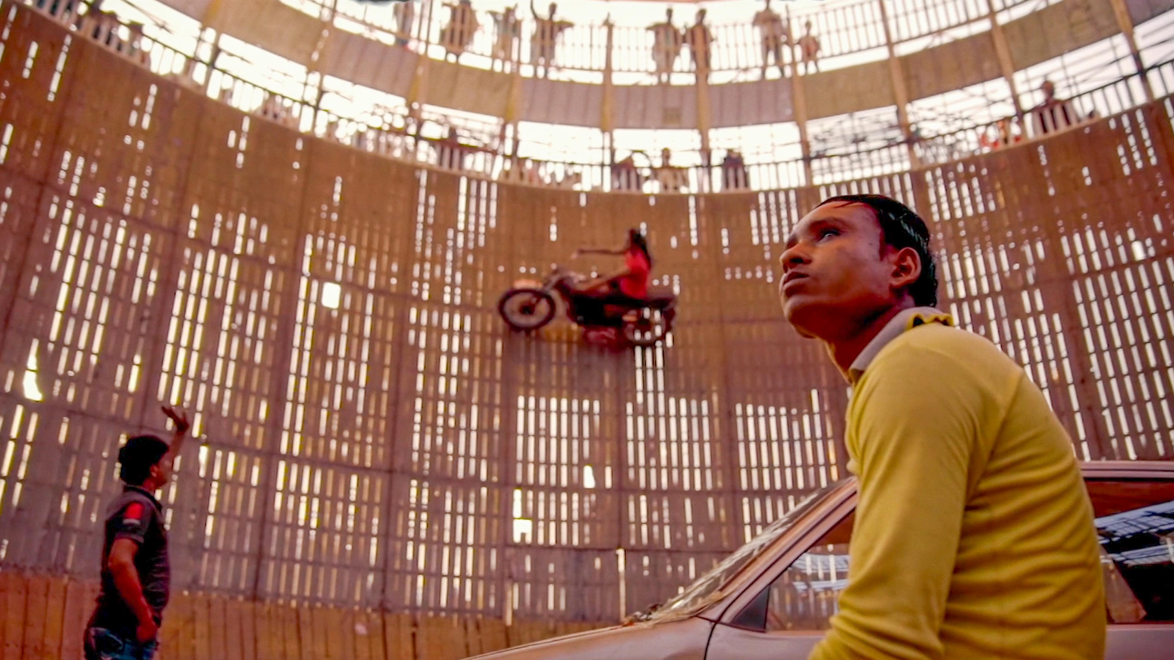 A person riding a motorcycle on a vertical wall in a wooden cylinder arena with spectators above, indoors.