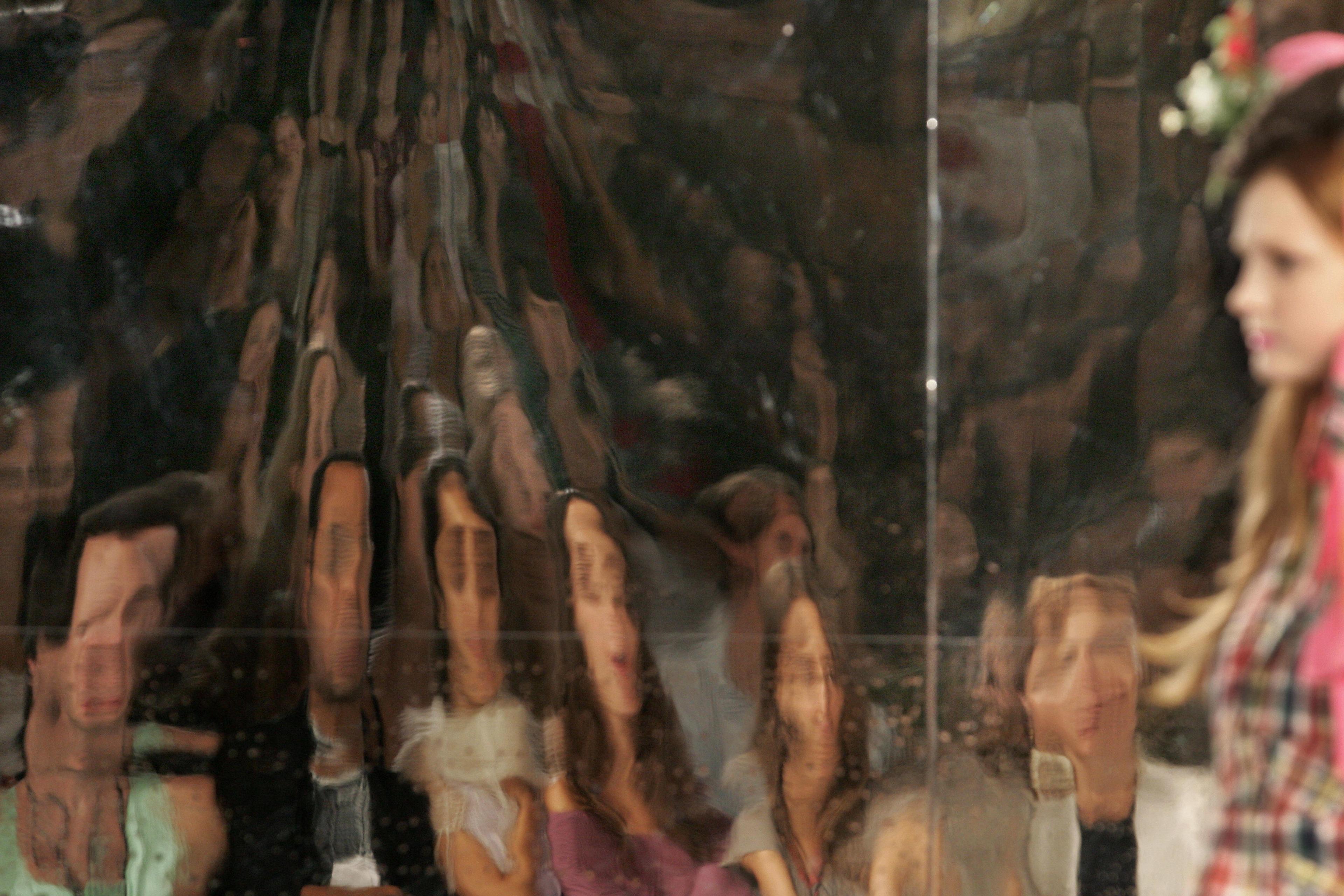 Distorted reflections of people on a textured surface next to a blurred person on the right, in a photo.
