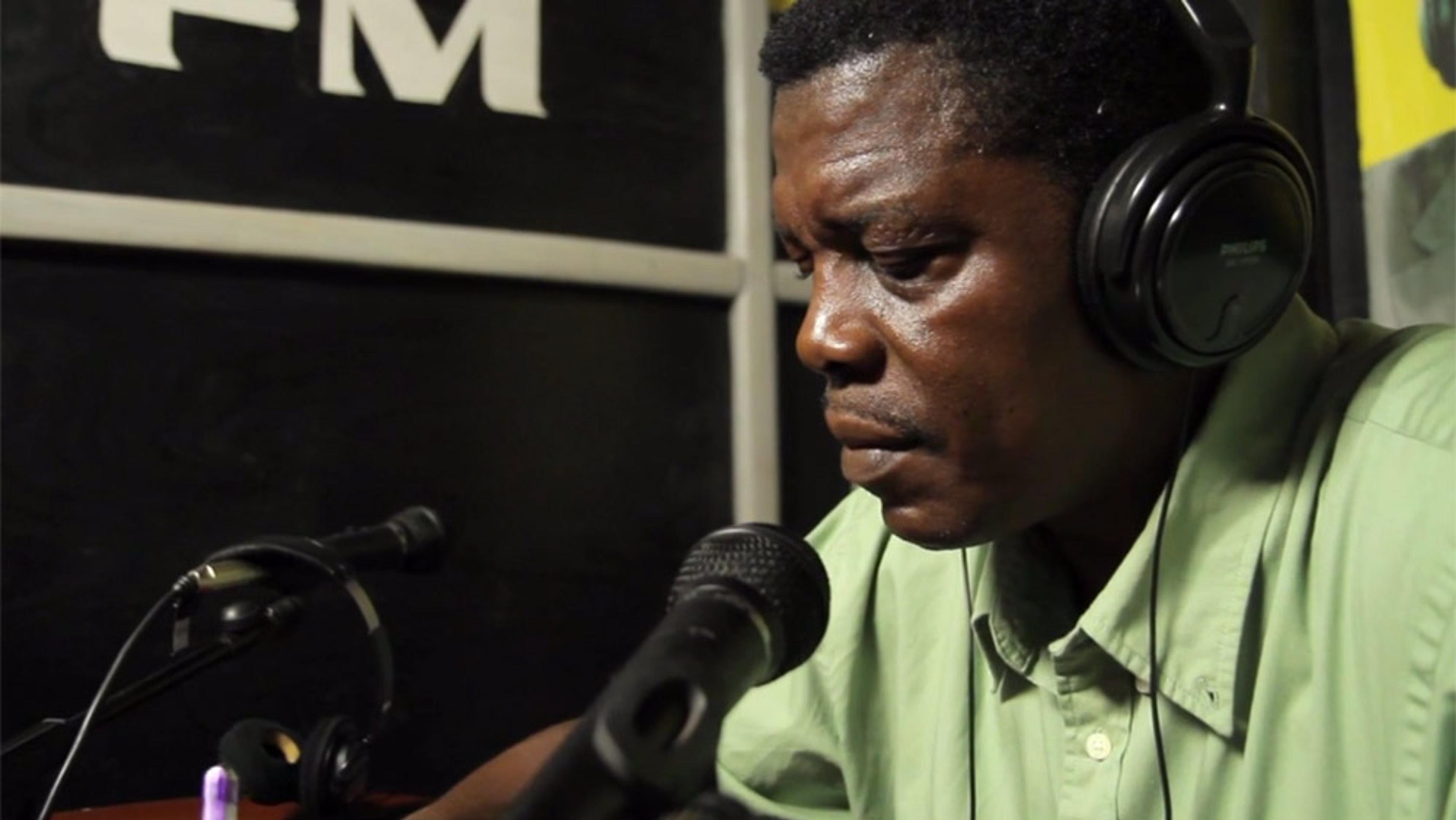 A man in a green shirt wearing headphones, speaking into a microphone in a radio studio with ‘FM’ visible in the background.