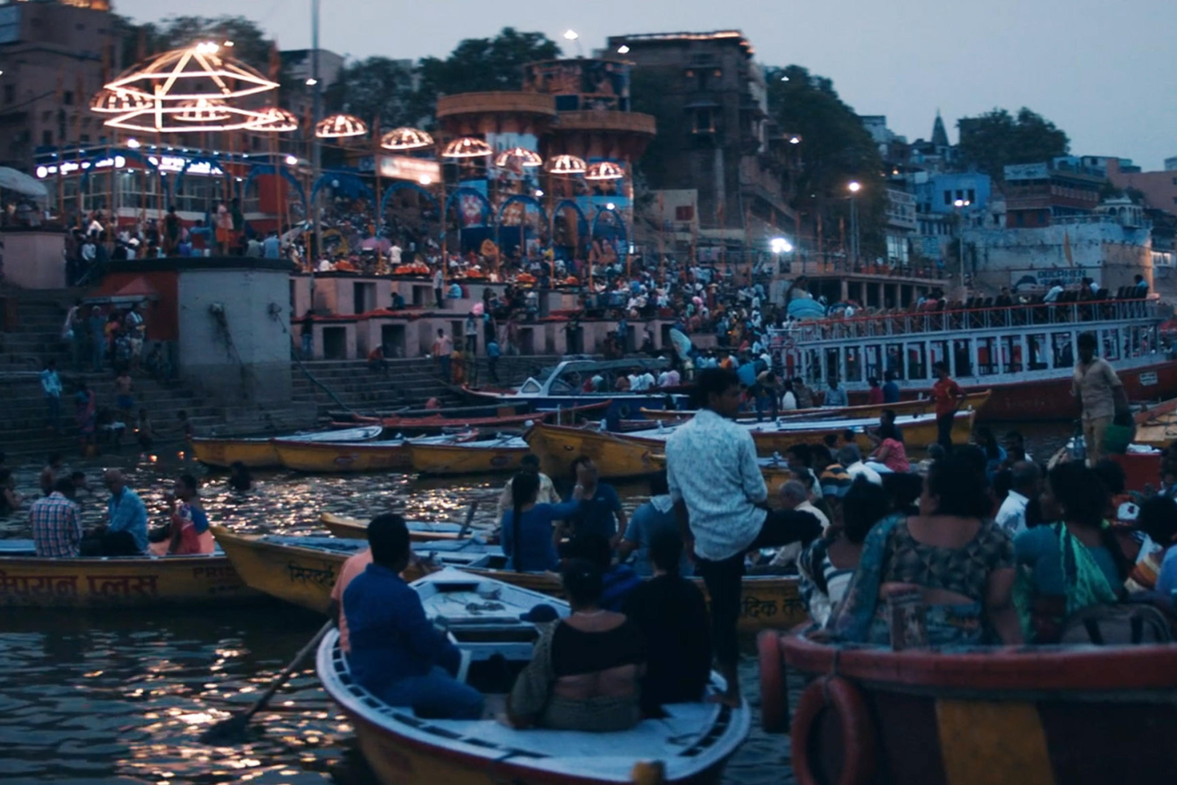 Crowded riverbank with numerous people in boats and on the ghats at dusk, celebrating a festival, lights in the background.