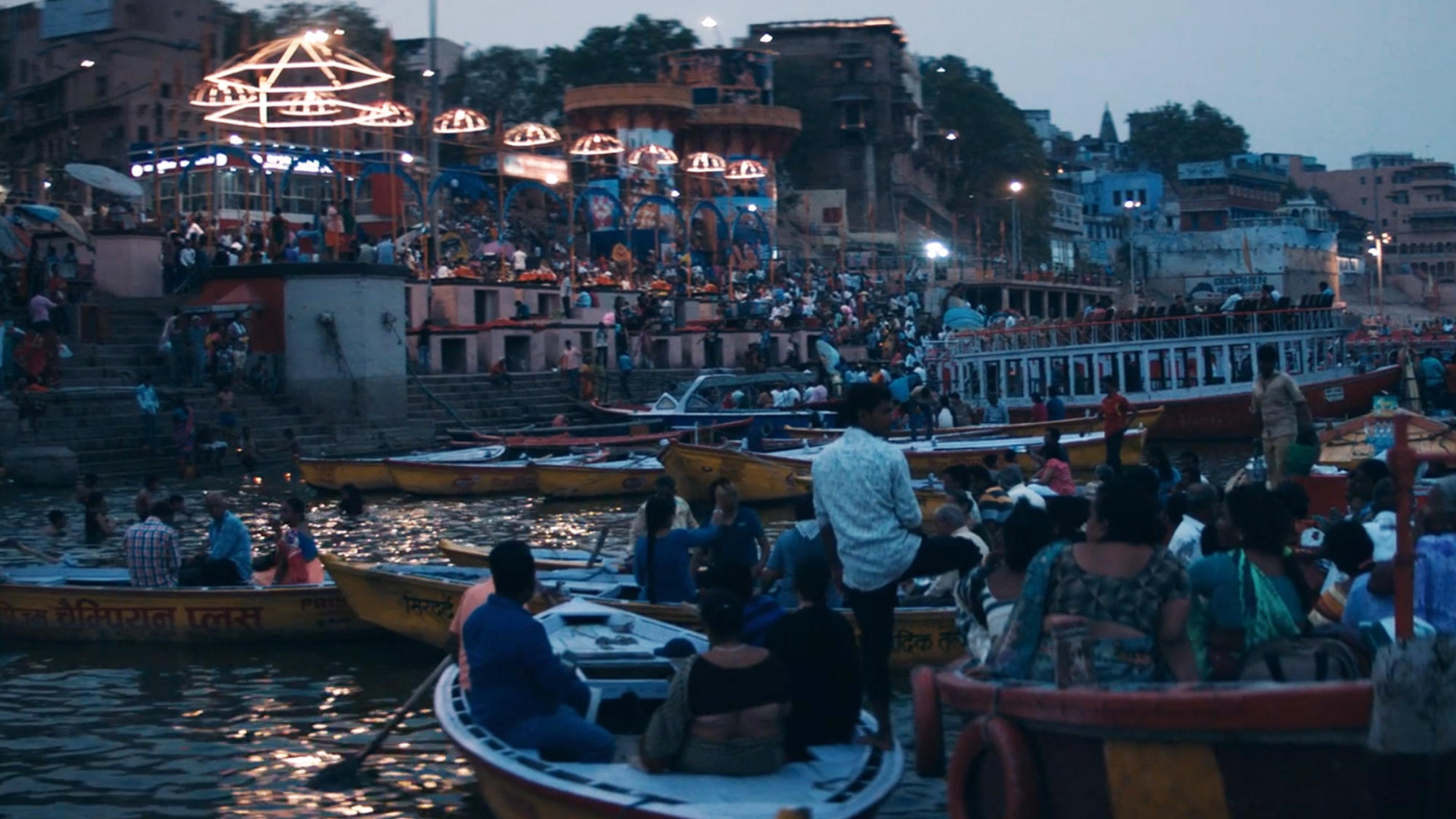 Crowded riverbank with numerous people in boats and on the ghats at dusk, celebrating a festival, lights in the background.