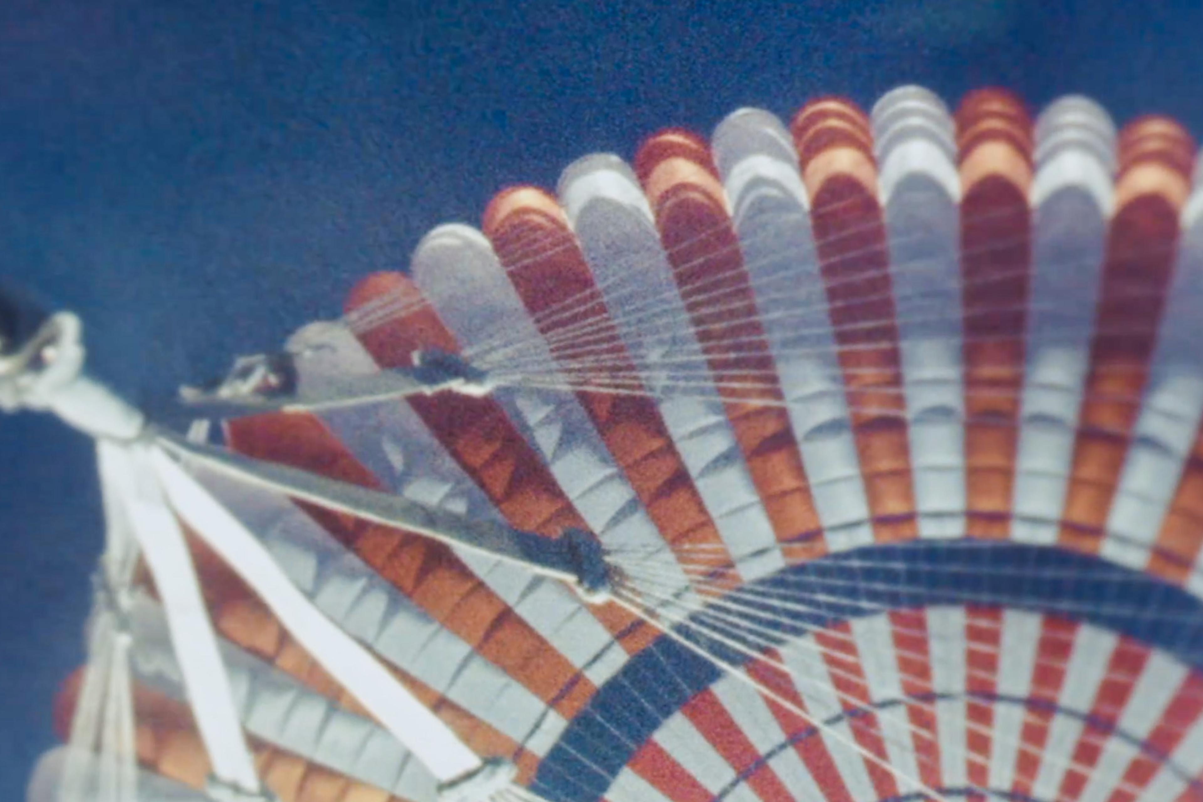 Close-up image of a NASA parachute with alternating orange and white panels against a clear blue sky.