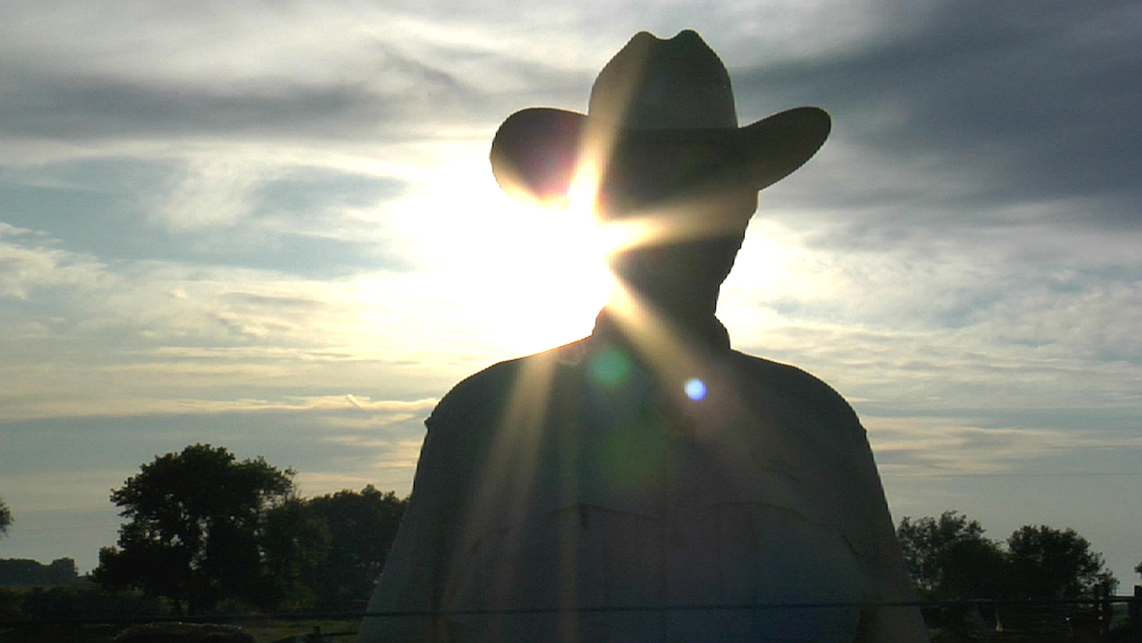 A man in a cowboy hat silhouetted against the sun with trees and a cloudy sky in the background.