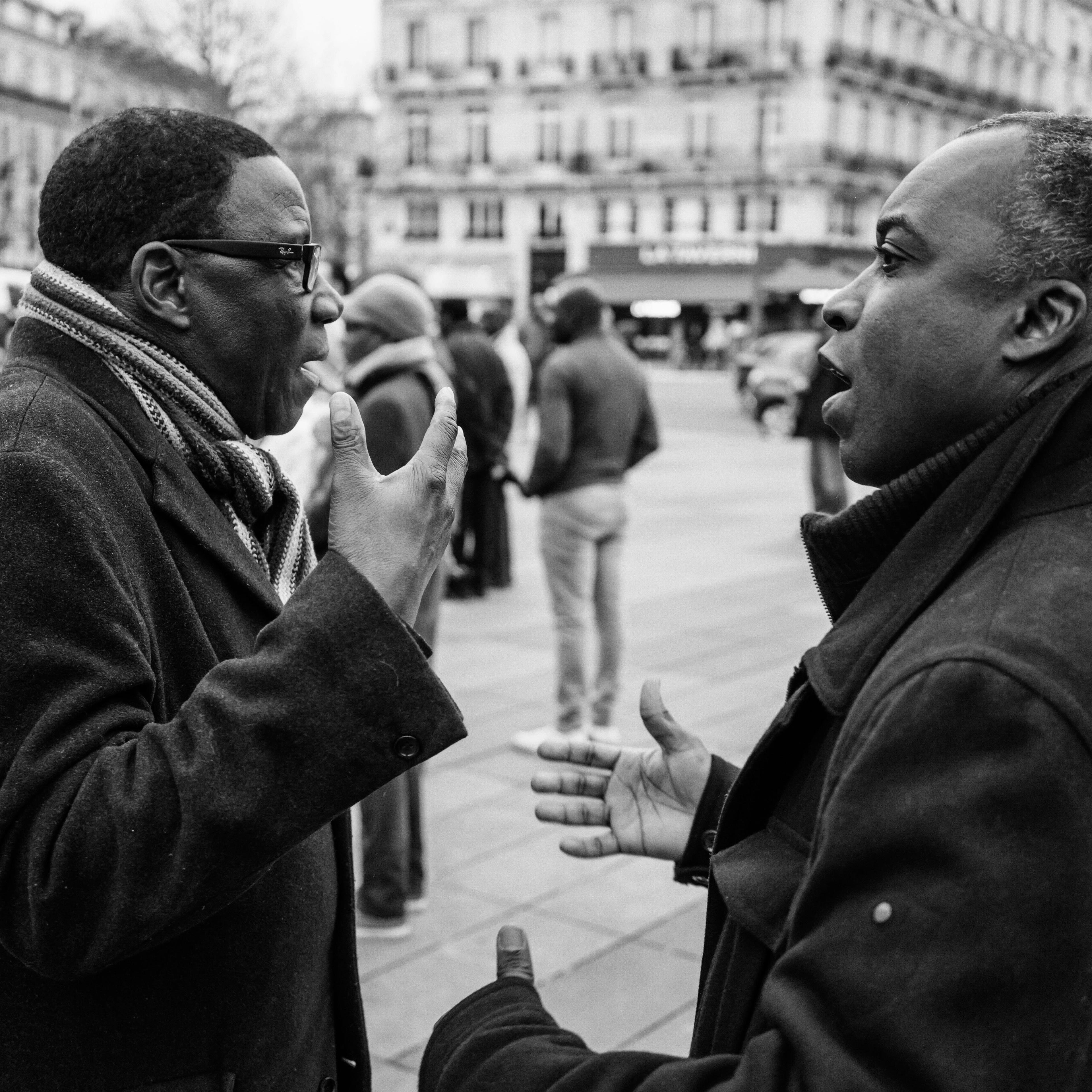 Black and white photo of two men in coats talking on a city street with blurred people and cars in the background.