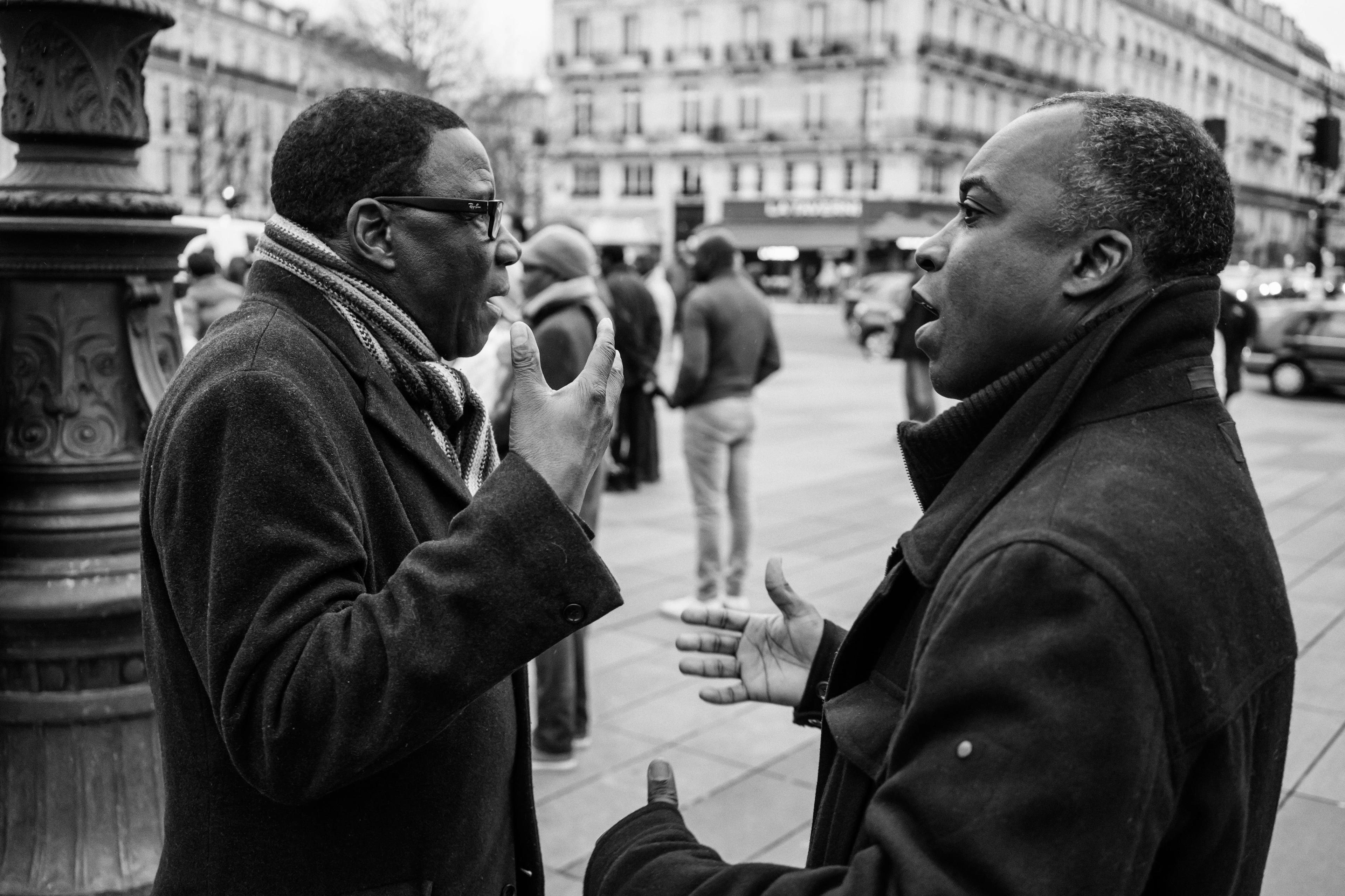 Black and white photo of two men in coats talking on a city street with blurred people and cars in the background.