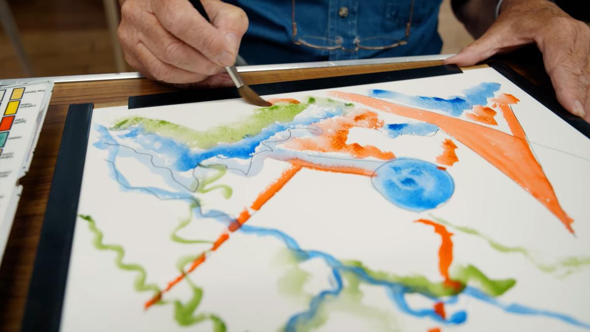 Person painting abstract watercolour with green blue orange shapes on paper, paint set visible on table.