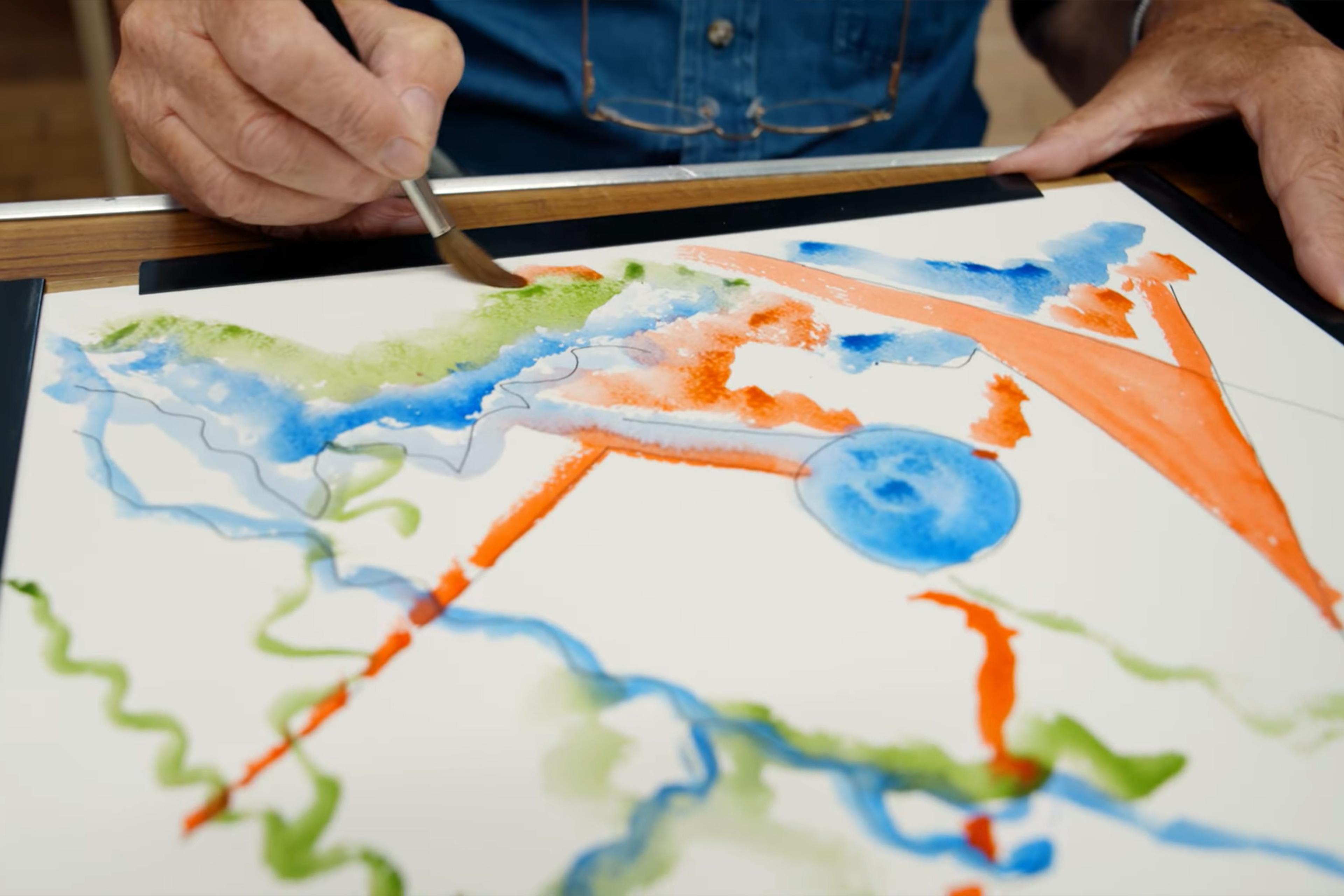 Person painting abstract watercolour with green blue orange shapes on paper, paint set visible on table.