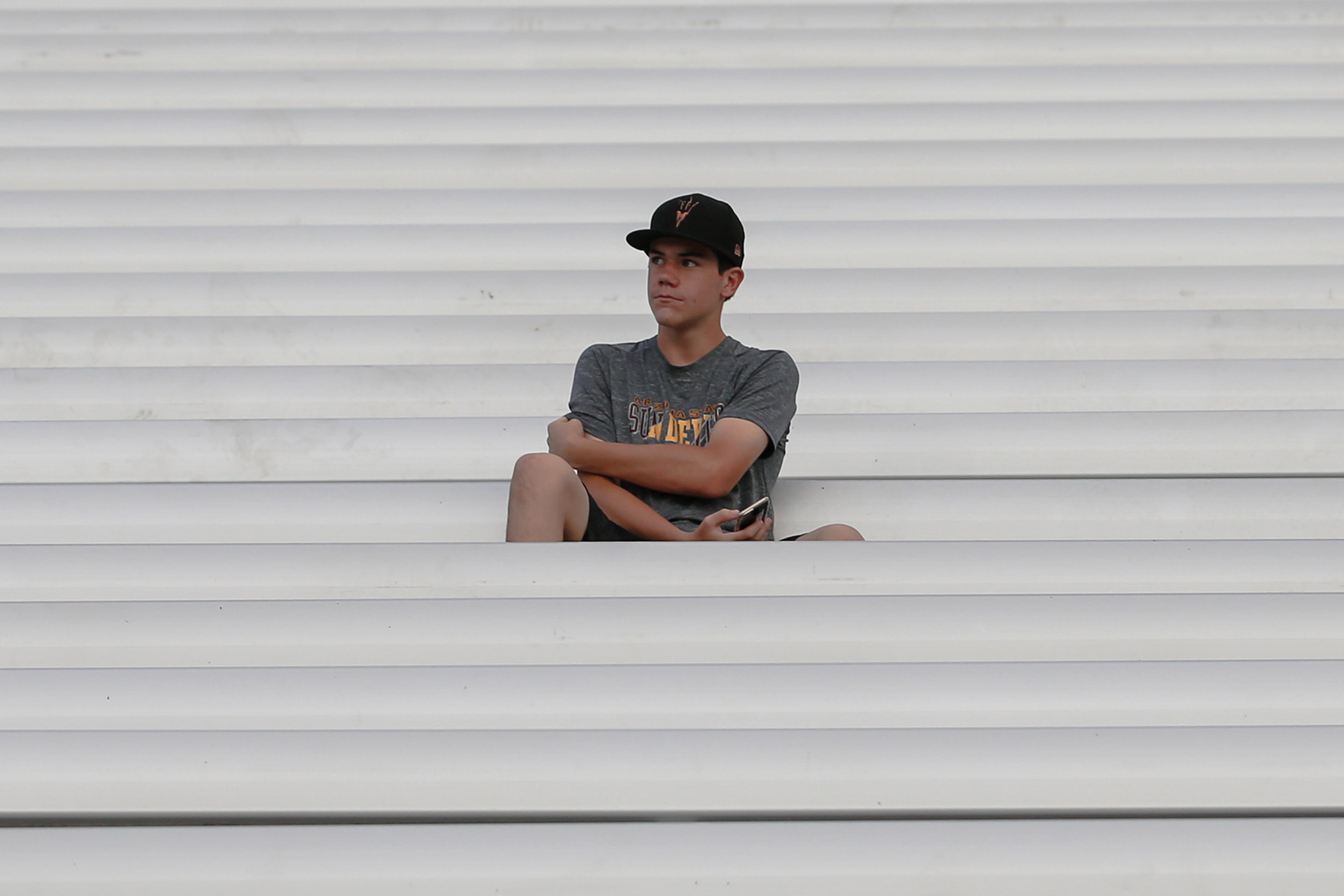 A boy wearing a cap and t-shirt sits alone on white stadium bleachers, looking to the side with his arms crossed.