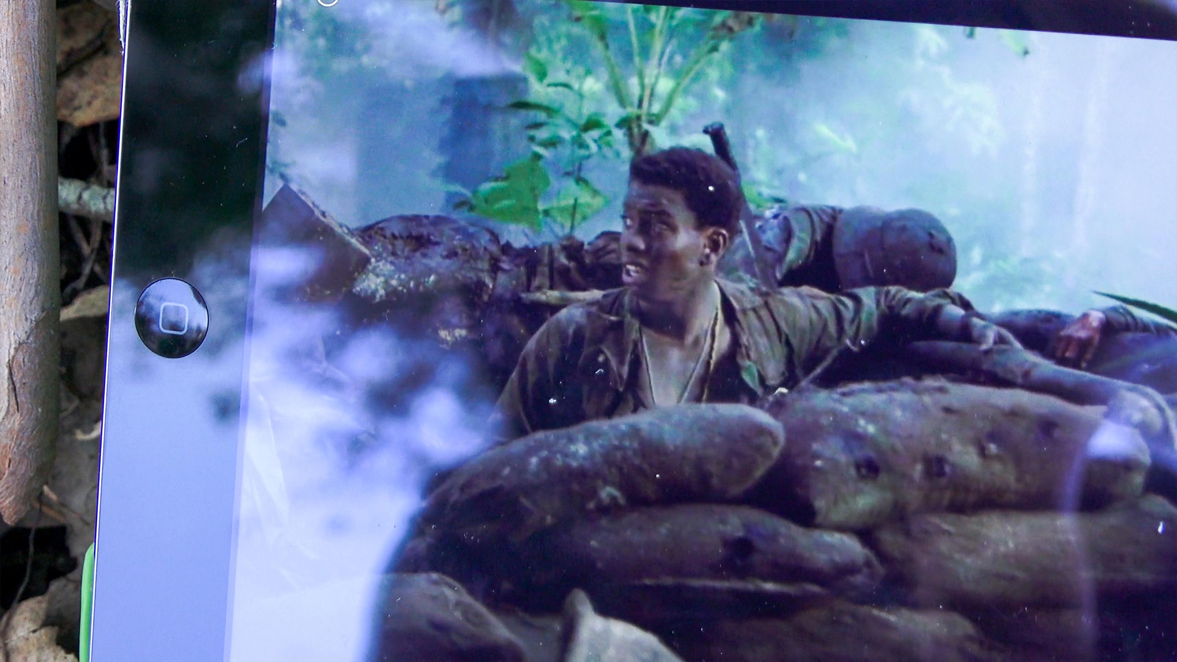 Photo of a scene from a film showing a soldier in muddy gear behind sandbags in a jungle, reflected on an iPad screen.
