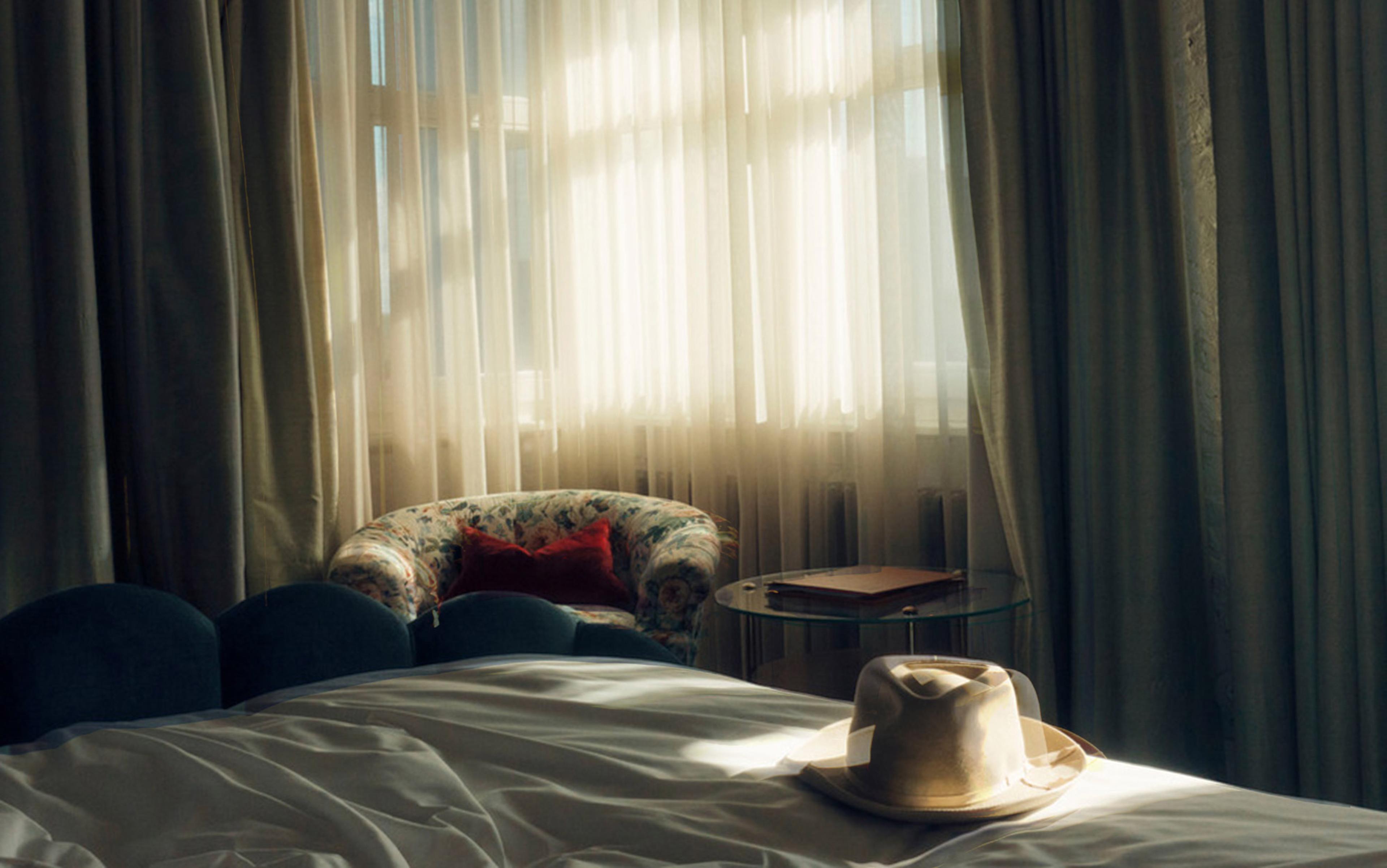 A Panama hat rests on a bed bathed in afternoon light filtering through gauze curtains. There is a slight motion blur