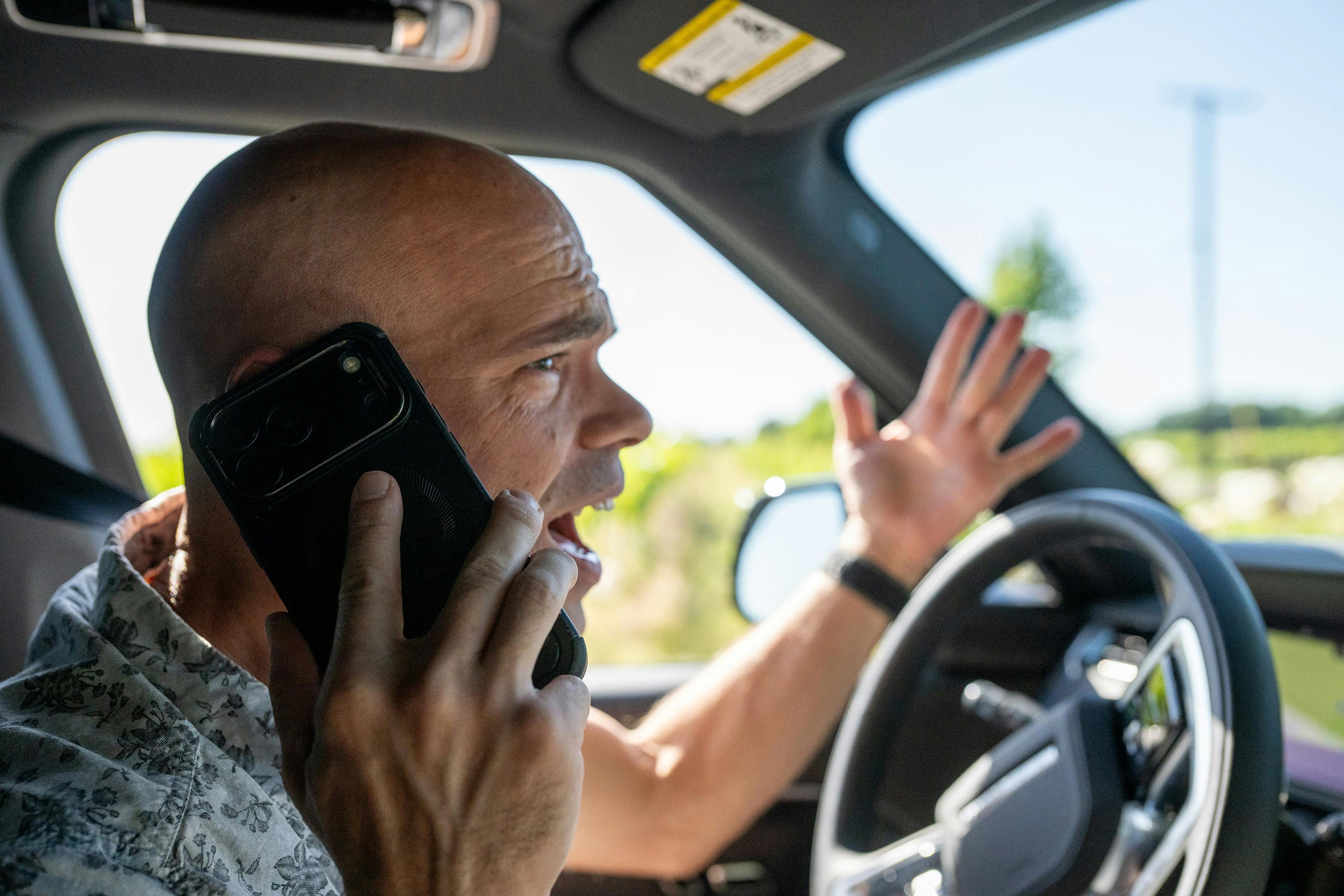 A man in a car talking on a phone and gesturing with his hand, appearing animated or upset.