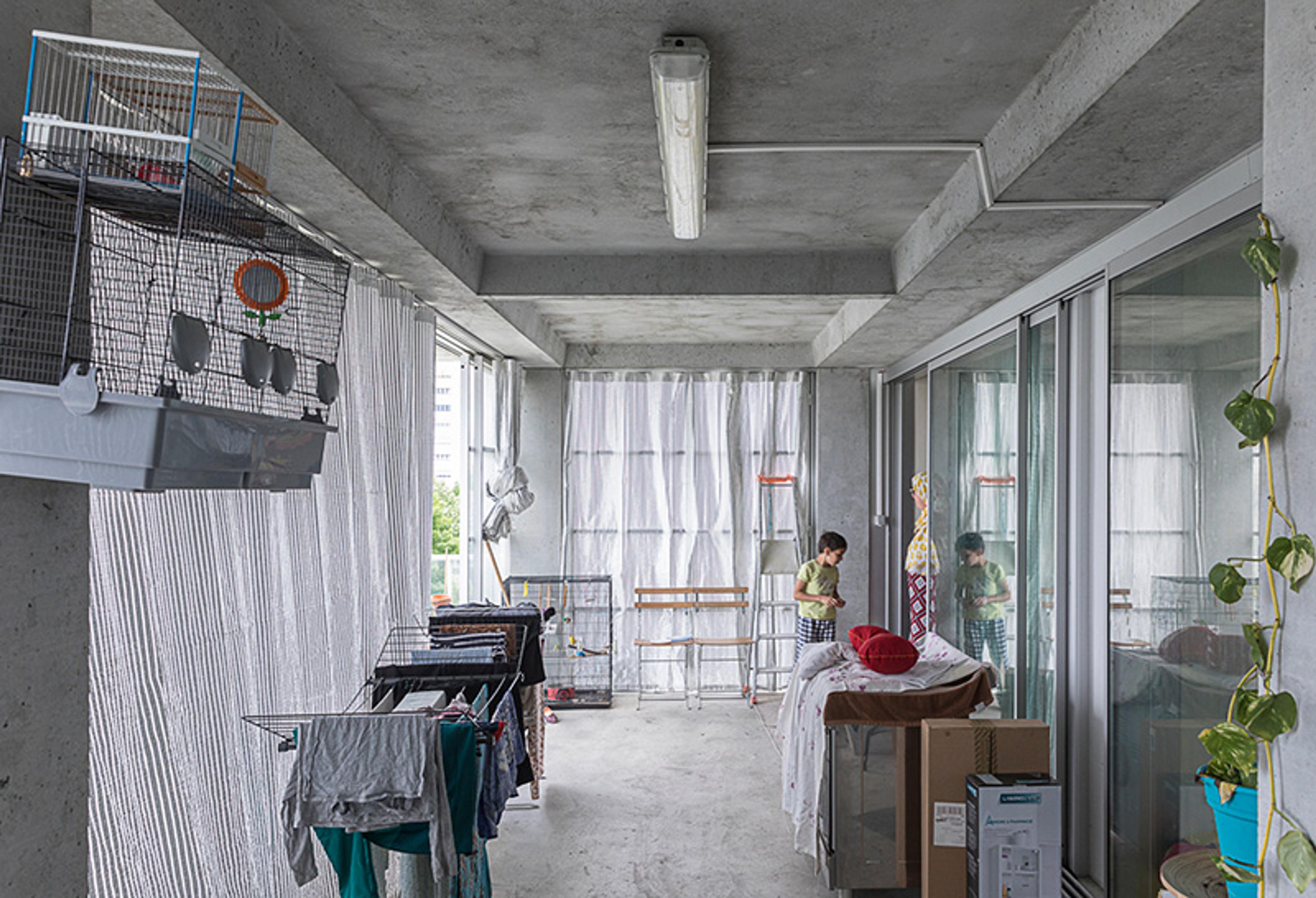 A concrete balcony with laundry drying racks, a birdcage, and a person in the background near a table and boxes.