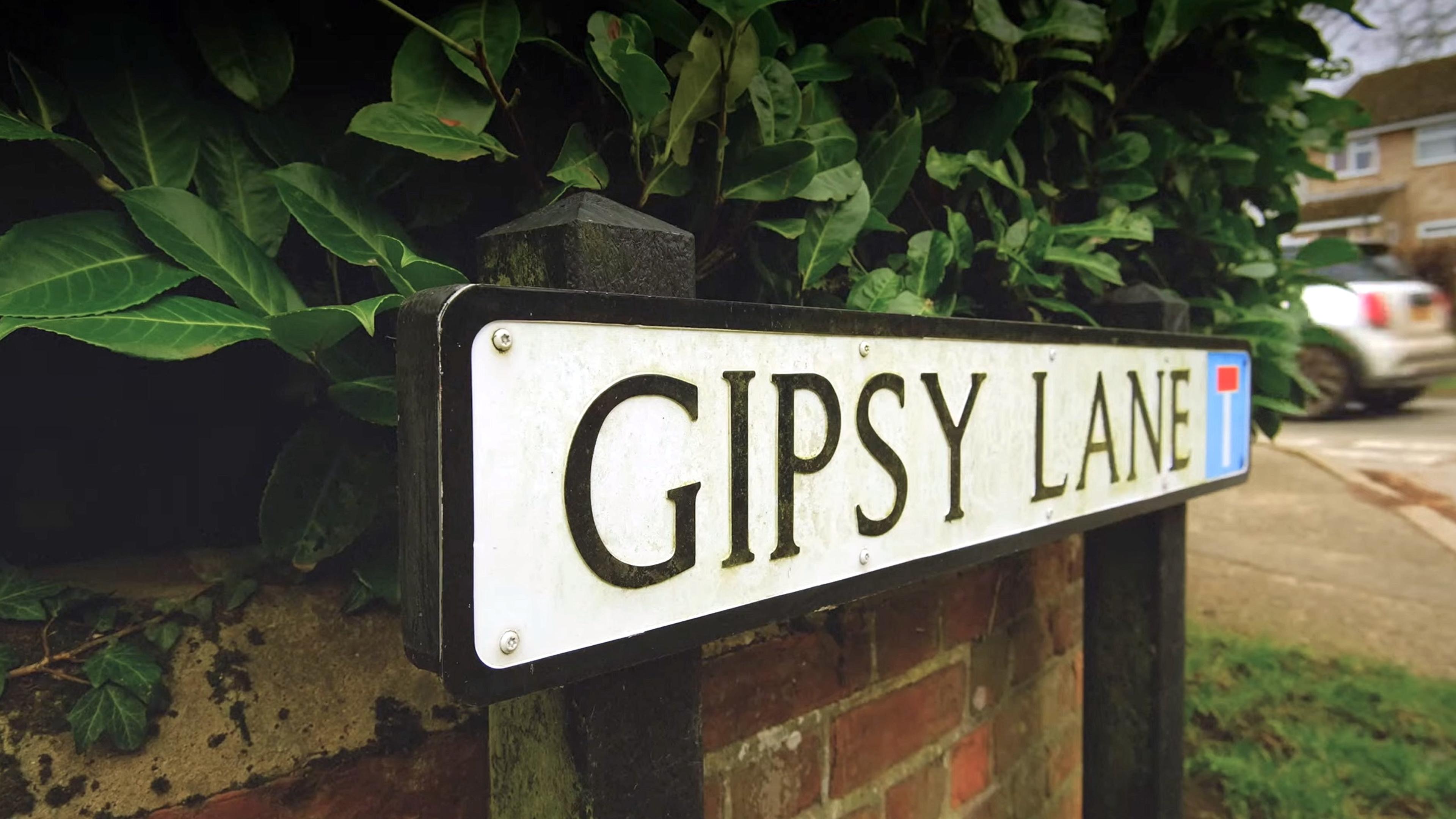 Photo of a street sign reading ‘Gipsy Lane’, mounted on a brick wall with green foliage behind, and a car in the background.